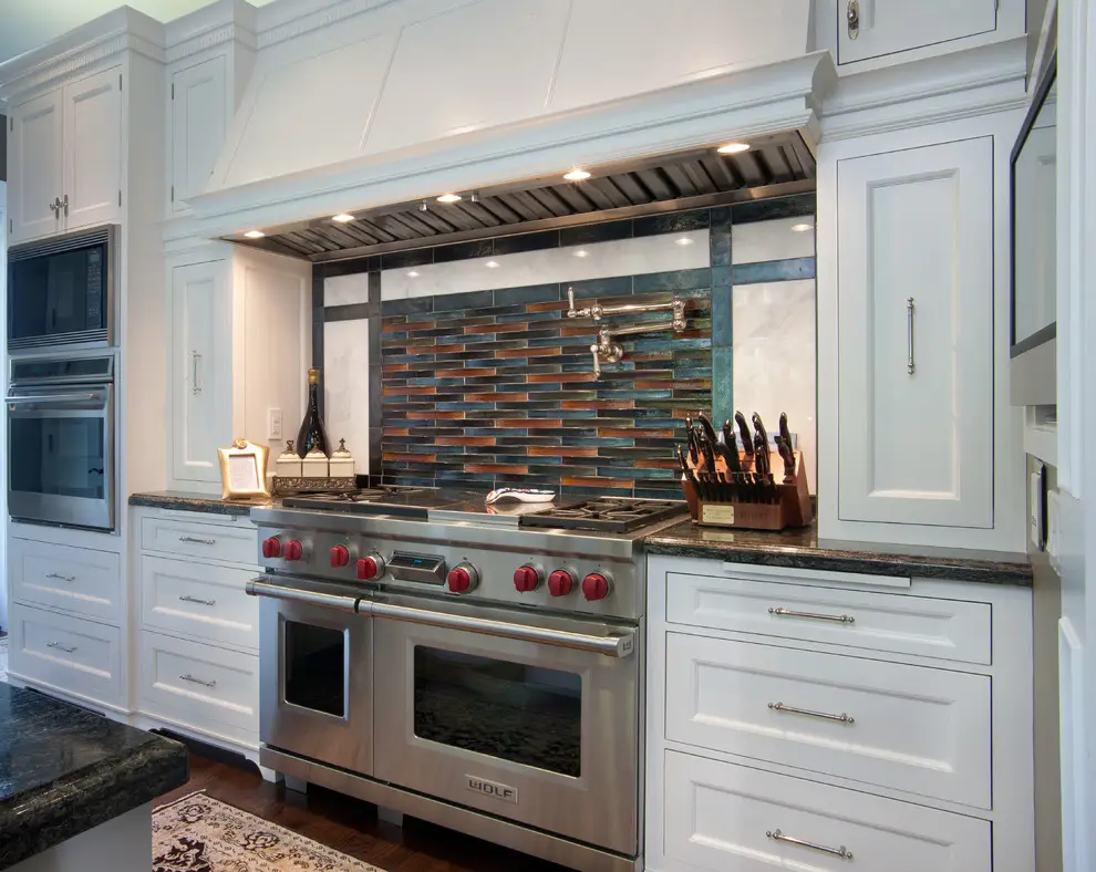 Modern kitchen featuring minimalist white cabinets and a stainless steel stove with red knobs. The backsplash displays a mix of blue and copper tiles, adding a sleek design element. The kitchen's organization enhances functionality and style.