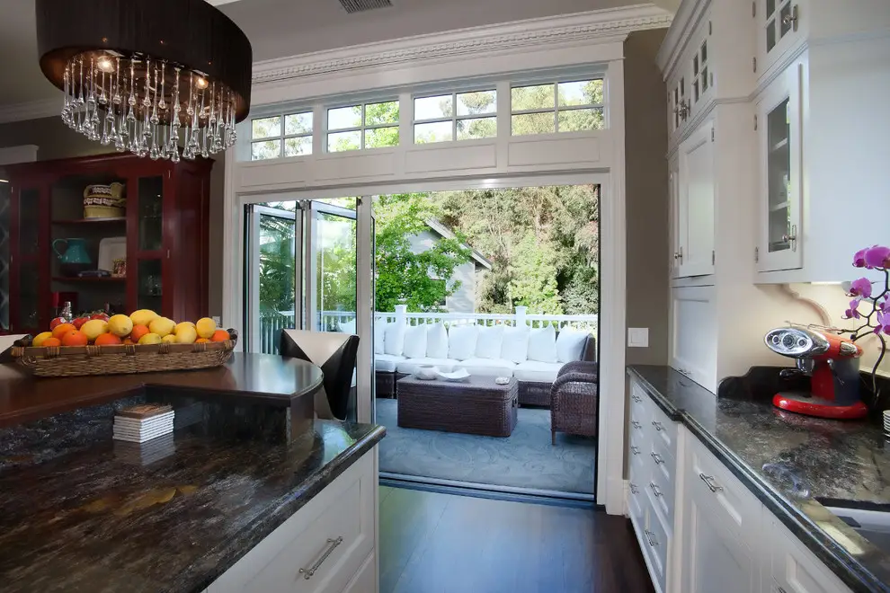 Modern kitchen with white cabinetry and dark countertops, featuring a view through open glass doors to a well-furnished patio. Highlights include a basket of fruits, a chandelier, and a red espresso machine, set in a minimalist design style.