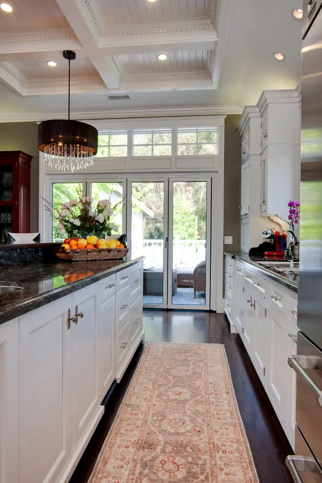 Sleek kitchen featuring modern and minimalist white cabinets with dark countertops. Elegant chandelier lighting complements the airy space with large windows and glass doors opening to outdoor seating, enhancing the contemporary ambiance.