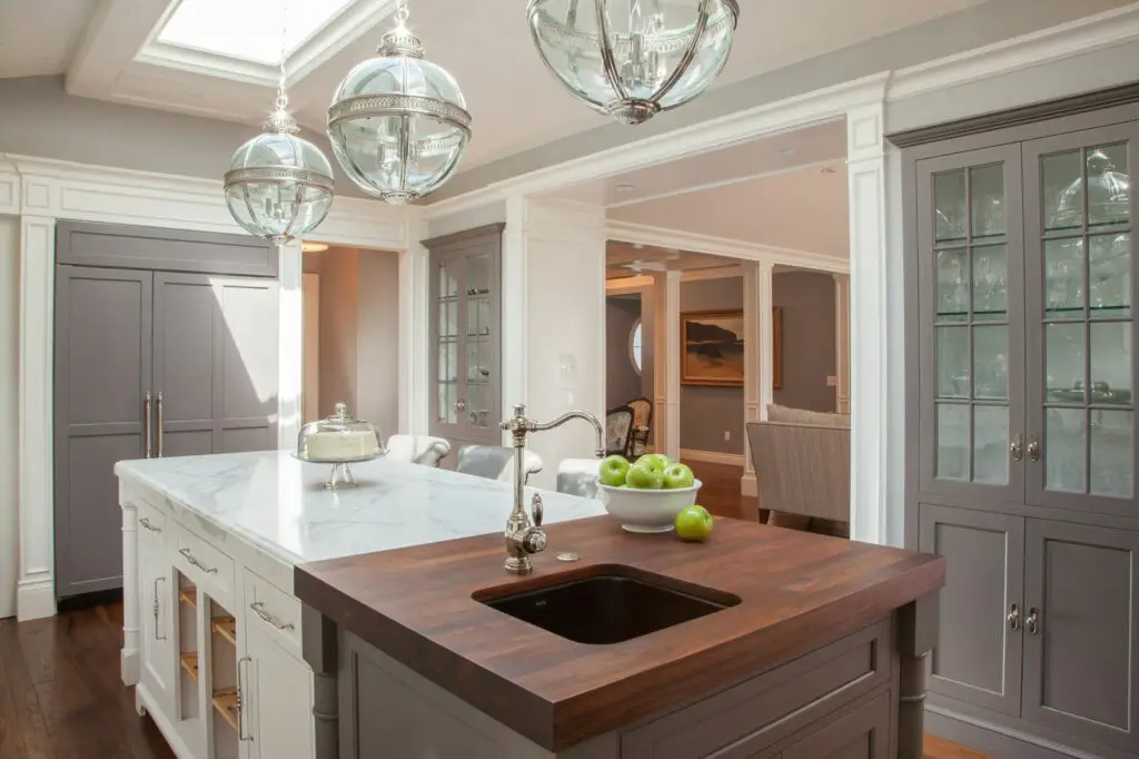 Luxurious kitchen with custom La Jolla cabinets featuring glass doors, marble countertops, and a wooden island with a sink, illuminated by elegant pendant lights; perfect elegant storage solution.