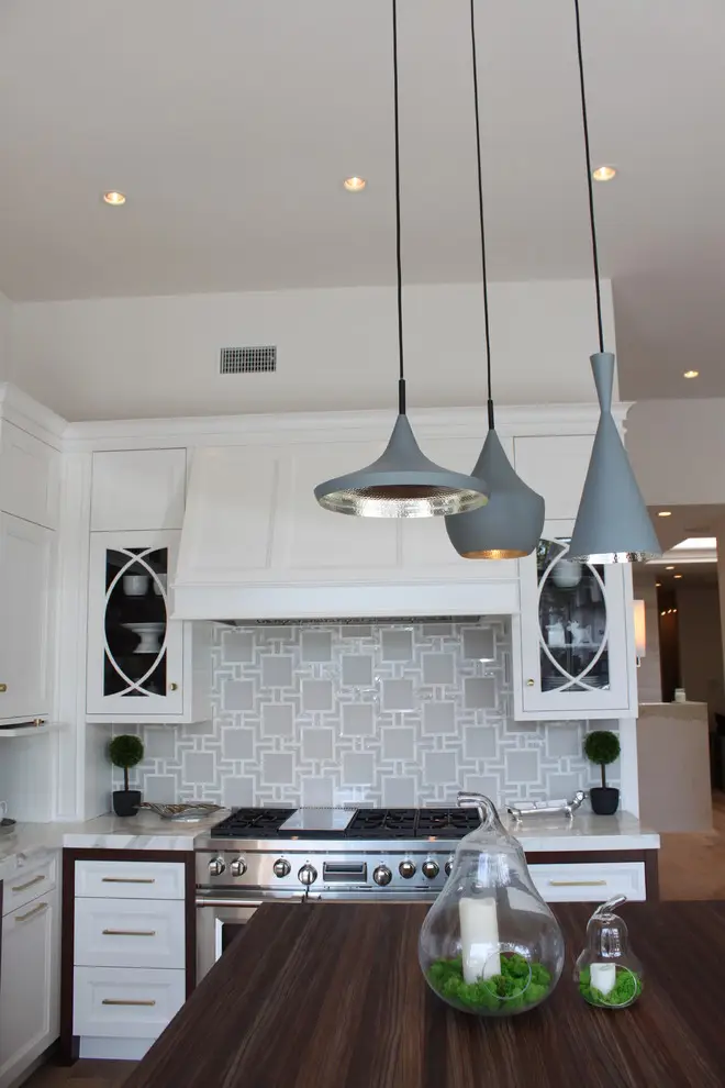 Modern kitchen with custom white cabinetry featuring geometric glass doors, elegant gray pendant lights, and a dark wooden countertop. A stainless steel stove is set against a patterned tile backsplash with stylish decor elements.