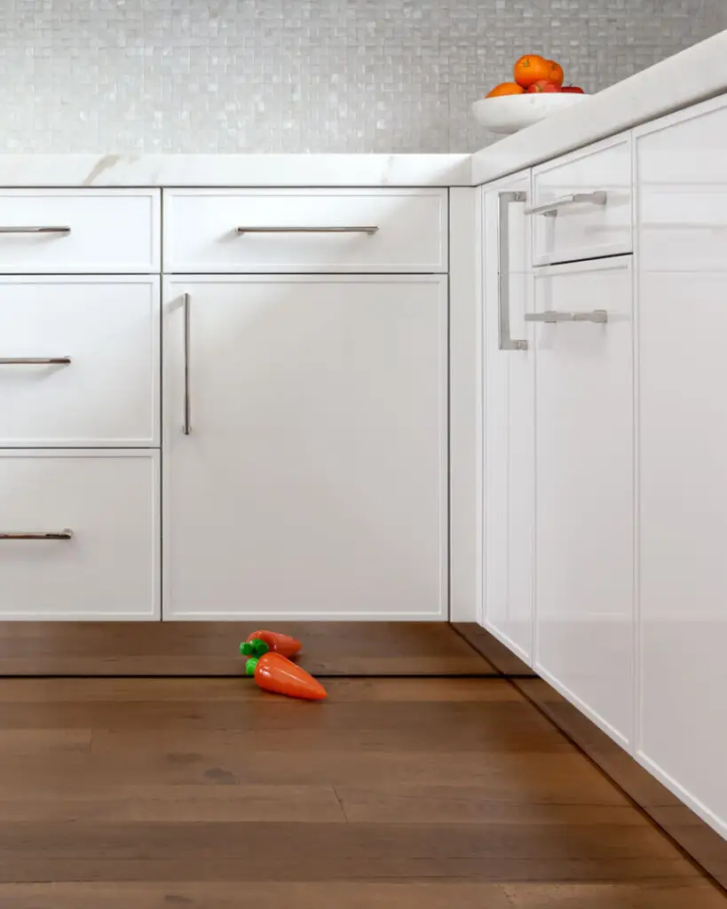 Modern kitchen interior with sleek white custom cabinetry, stainless steel handles, and a marble countertop. Wooden flooring enhances the elegant design. A bowl of fruit is placed on the counter, complementing the overall premium look of the kitchen in a La Jolla interior design studio.