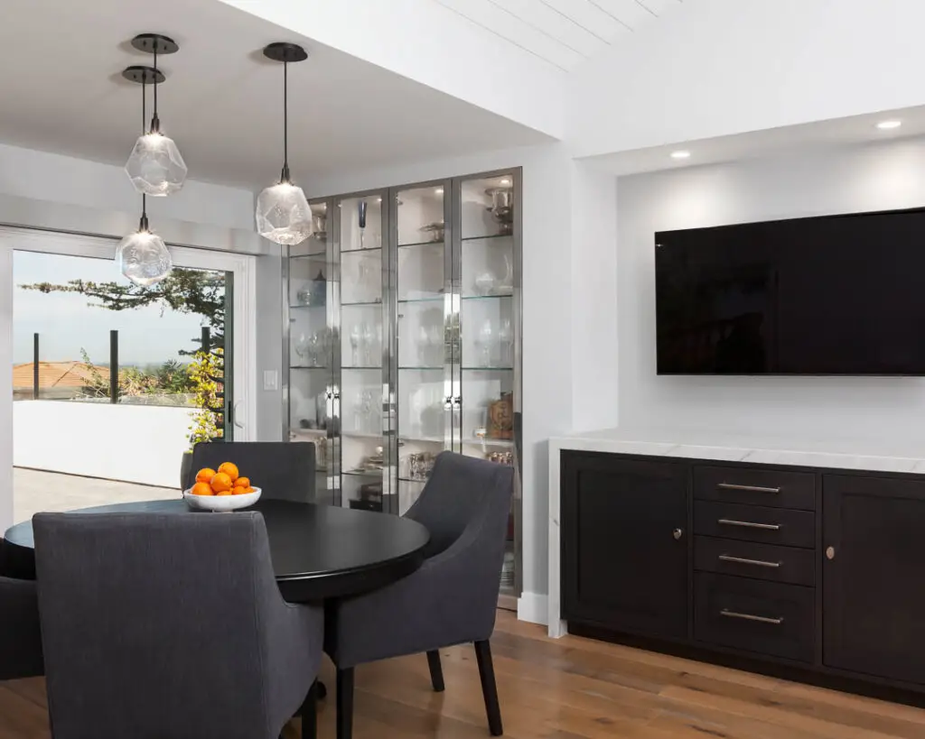 Modern dining room with custom cabinetry in La Jolla interior design studio, featuring a round table with dark upholstered chairs, pendant lighting, and a glass cabinet showcasing elegant glassware.
