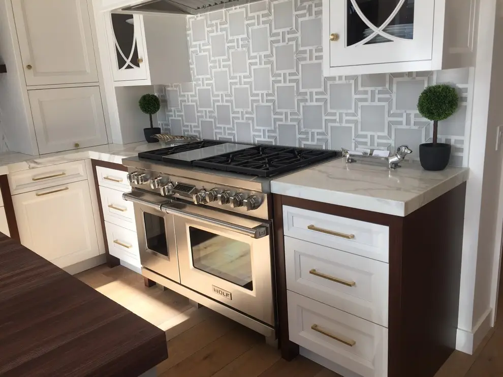Modern kitchen with custom white cabinetry featuring gold handles, a stainless steel stovetop and oven, and a geometric patterned backsplash. Elegant storage design includes decorative plants on marble countertops.