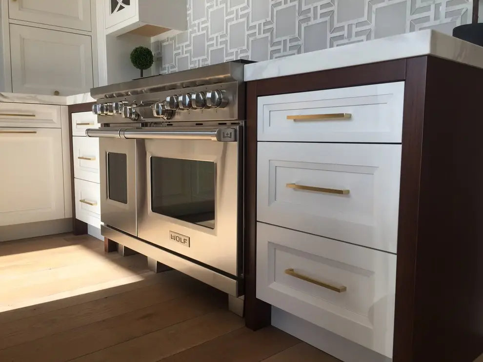 Custom cabinetry with sleek white drawers and golden handles in a modern kitchen, featuring a stainless steel stove and geometric tile backsplash, emphasizing elegant storage solutions.