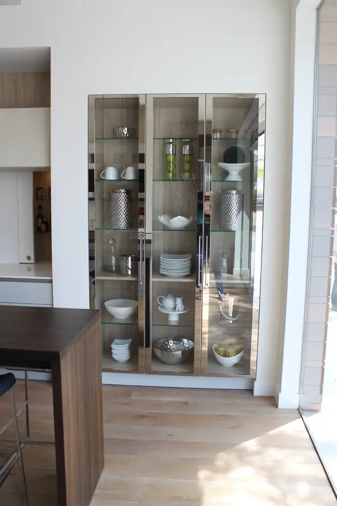 Glass-front custom cabinetry in a modern kitchen design featuring elegant storage for dishes and decorative items, with a wooden countertop and light wood flooring providing a contemporary aesthetic.