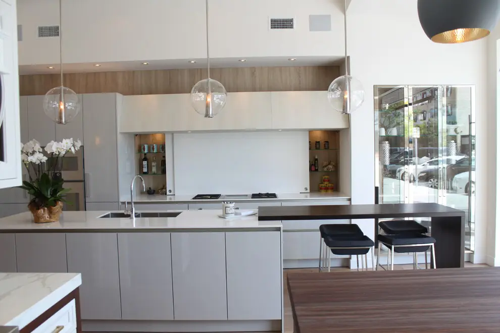 Modern kitchen with custom cabinetry, featuring sleek white cabinets, built-in appliances, and a central island with dark wood accents. Elegant pendant lights and a vase with white flowers add sophistication. Large windows allow natural light to enhance the stylish design.