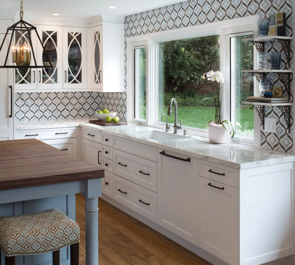 Luxury custom kitchen cabinetry and organizers in Los Angeles, featuring white cabinets with geometric glass inserts, a spacious countertop, and modern hardware. The kitchen includes a patterned backsplash, ample natural light from large windows, and an elegant pendant light over a wooden table.