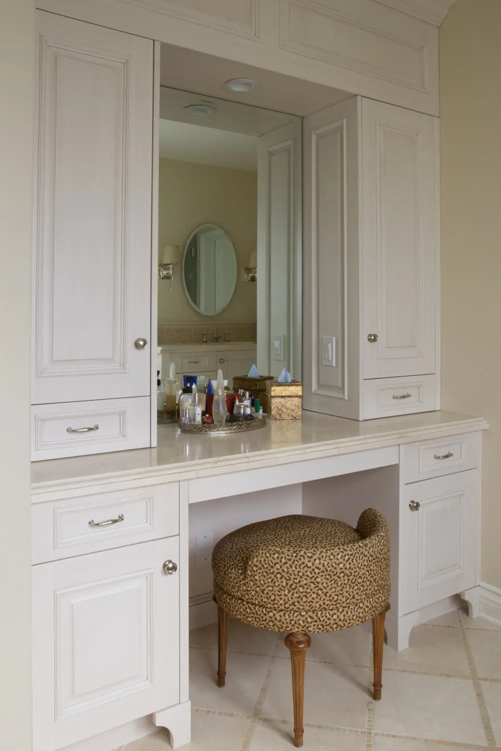 Elegant custom cabinetry with a beige finish, featuring built-in drawers and cabinets surrounding a mirror. The countertop displays various toiletries. An upholstered wooden stool with a leopard print cushion is tuc