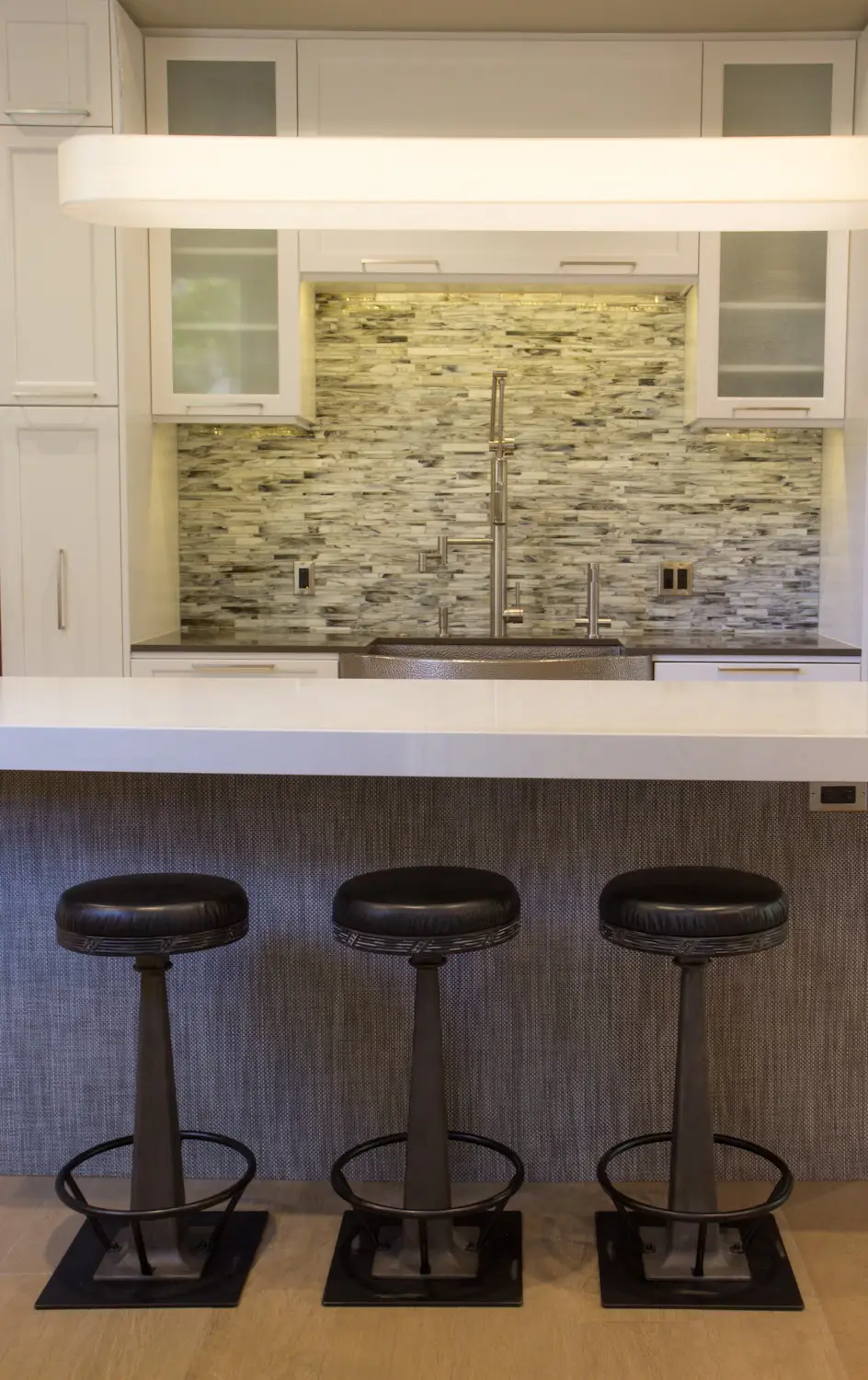 Modern kitchen with custom cabinetry, featuring a sleek design with a marble backsplash, white cabinets, and a white countertop. Includes three black leather bar stools and a contemporary faucet, highlighting efficient storage solutions in a stylish setting.