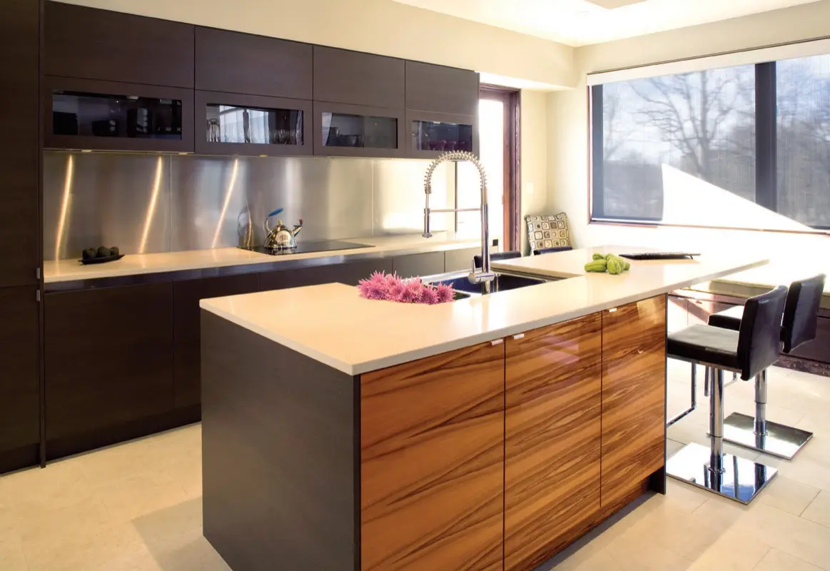Modern kitchen design featuring custom cabinets and an island with a sleek countertop, stainless steel backsplash, and contemporary faucet. Large windows allow natural light, enhancing the elegant and stylish atmosphere.