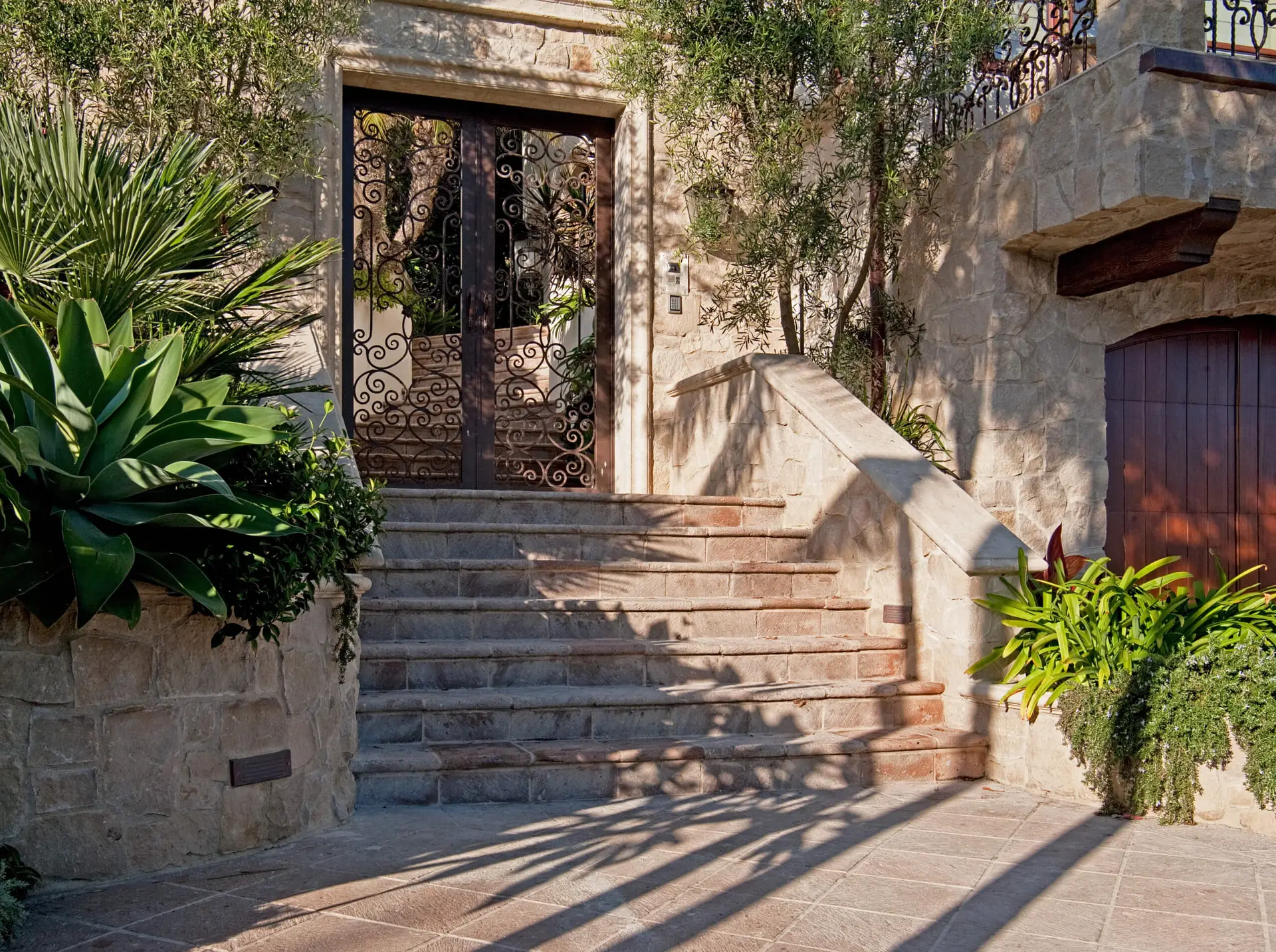 Elegant stone staircase leading to a wrought iron gate, surrounded by lush green plants, showcasing an inviting entrance to a luxurious modern home.
