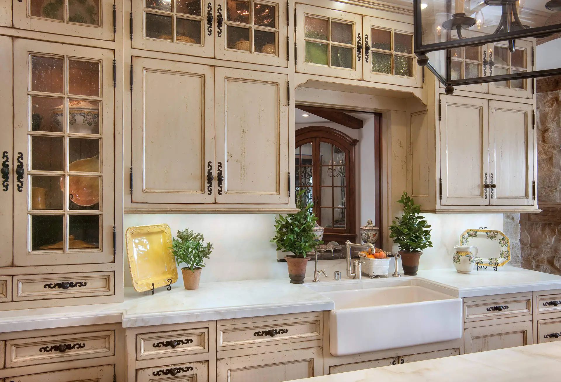 Luxury kitchen design featuring custom cabinetry with glass-front doors and vintage handles, marble countertops, and farmhouse sink, decorated with potted plants and elegant kitchenware for modern homes.