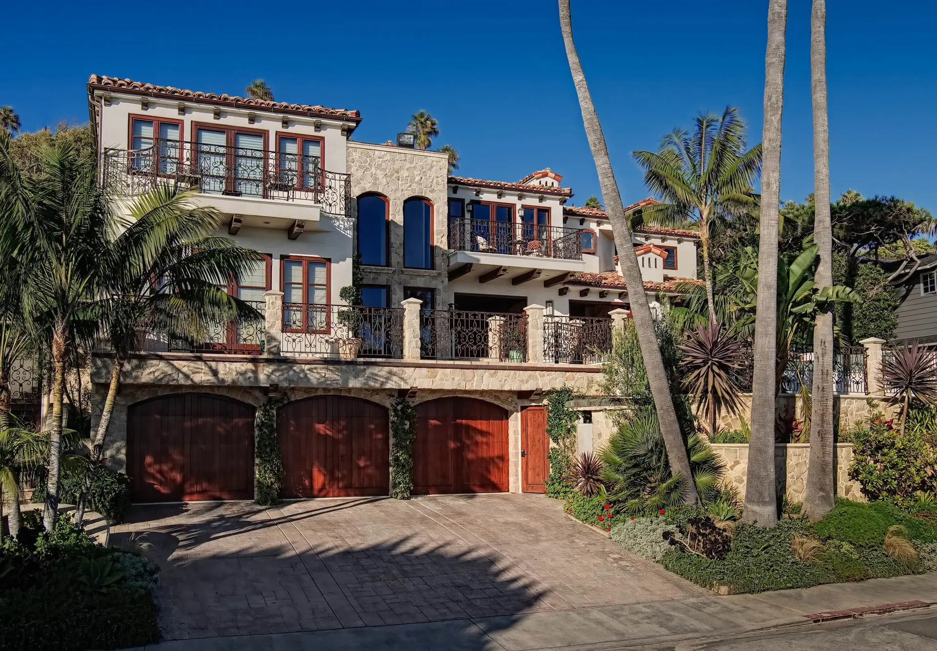 Front view of a luxurious Mediterranean-style home with custom design, featuring stucco walls, red tile roof, and wrought iron balconies, surrounded by palm trees and lush landscaping.