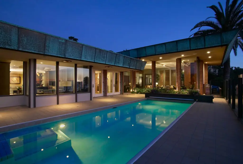 Modern home exterior with illuminated interior viewed through large windows, featuring a sleek pool surrounded by tiled patio under evening sky.