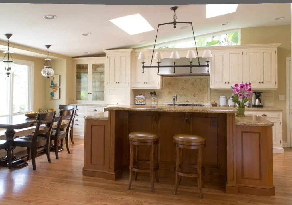 Luxury kitchen design featuring custom white cabinetry, granite countertops, and elegant light fixtures. A wooden island with matching bar stools adds warmth, while a dining area with a large wooden table provides space for elegant dining. Skylights enhance the bright, open feel, complementing the sophisticated design of the kitchen space.
