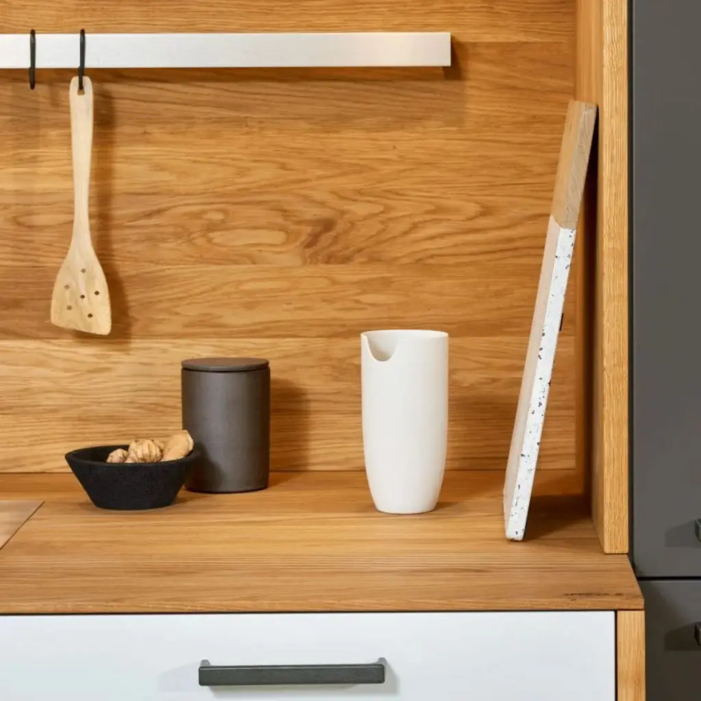 Modern kitchen cabinetry featuring a wooden countertop with a light wooden spatula hanging from a rail, a black bowl with ginger, a dark container, a white ceramic pitcher, and a cutting bo