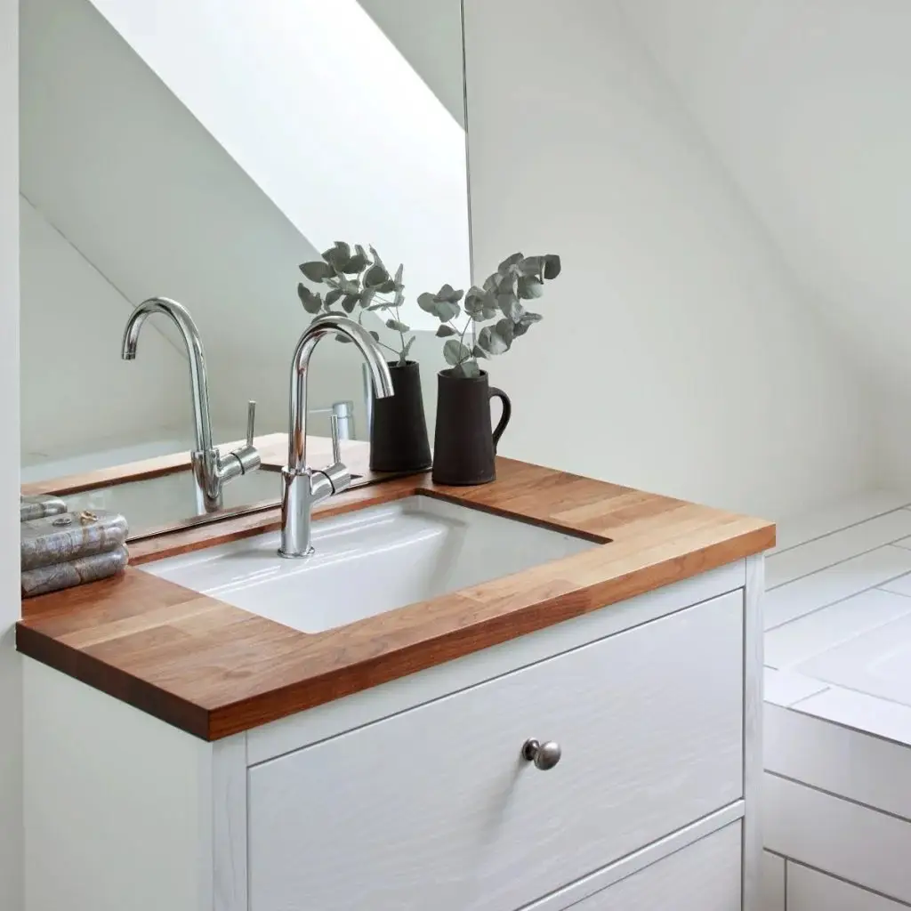Modern bathroom sink with a sleek chrome faucet and a wooden countertop. A large mirror reflects the minimalist design, complemented by decorative plants. Ideal for custom cabinetry inspiration at Inplace Studio.