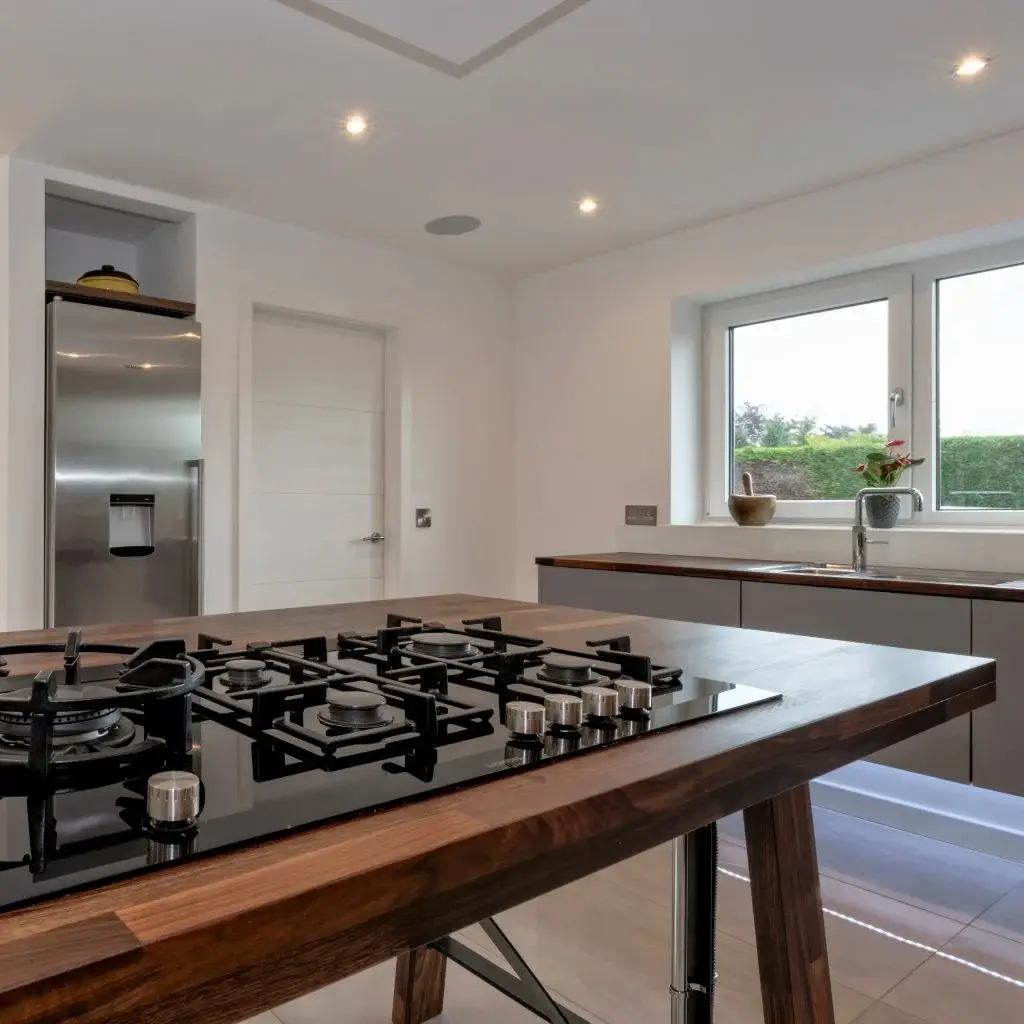 Modern kitchen interior featuring sleek black gas stovetop on wooden island, stainless steel refrigerator, and large windows providing natural light. Custom cabinetry offers ample storage space, exemplifying contemporary kitchen design by Inplace Studio.