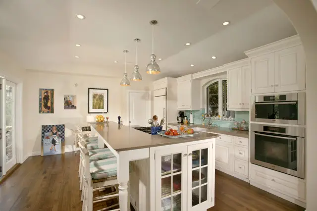 Spacious transitional and modern kitchen with white cabinetry and a central island. The kitchen features stainless steel appliances, pendant lighting, and a mix of contemporary and traditional decor elements. A hardwood floor complements the light color scheme, creating a bright and welcoming atm
