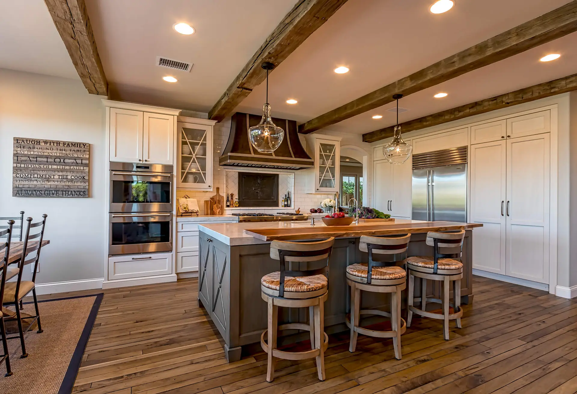 High-end European-style kitchen in Los Angeles featuring custom cabinetry. The kitchen showcases wooden beams on the ceiling, a large island with bar stools, a double oven, a stainless steel refrigerator, and elegant pendant lighting. The d&eacute;cor includes a mix of modern and rustic elements, creating a warm and inviting atmosphere.