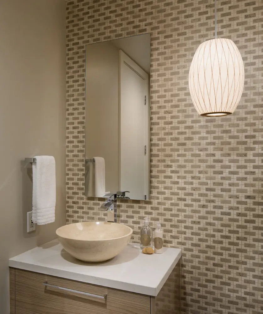 Elegant bathroom design featuring a beige stone vessel sink, modern faucet, and stylish geometric wall tiles, complemented by a light wood vanity and contemporary pendant light.