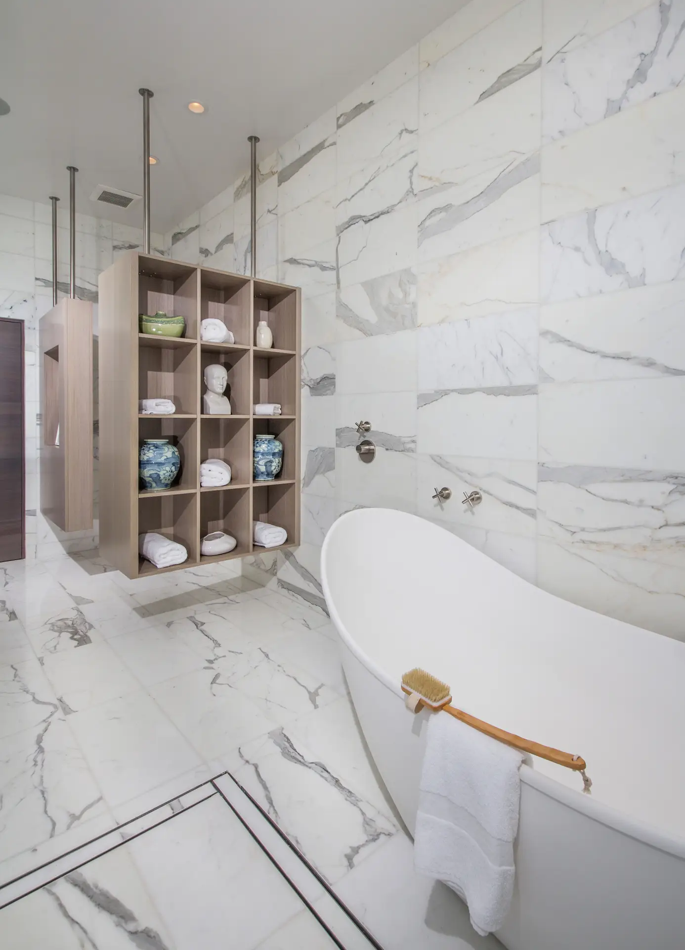 Luxury bathroom with marble tiles, featuring a modern freestanding bathtub and custom wooden shelving with decorative items and towels.