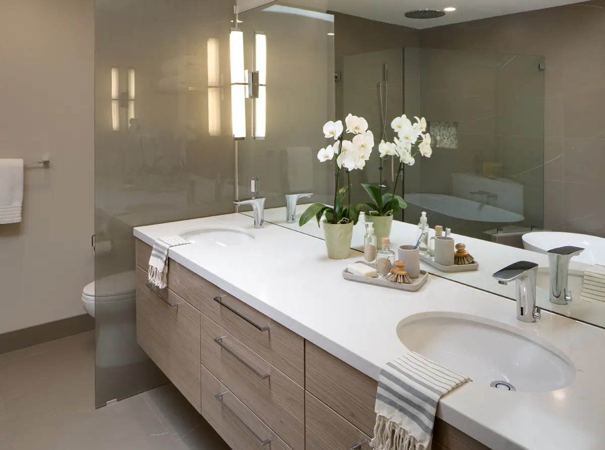Modern bathroom design featuring a luxury vanity with custom wood cabinets, sleek double sinks, and stylish chrome faucets. The white countertop is adorned with elegant orchids and stylish bath accessories, enhancing the contemporary feel.