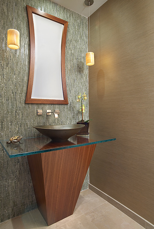 Modern bathroom design featuring a triangular wooden vanity with a glass countertop and a unique vessel sink. The room is completed with a textured tile backsplash, a contemporary mirror with a wooden frame, and pendant lighting.