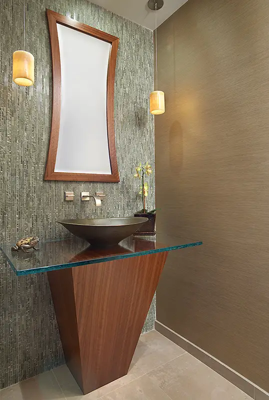 Modern bathroom design featuring a triangular wooden vanity with a glass countertop and a unique vessel sink. The room is completed with a textured tile backsplash, a contemporary mirror with a wooden frame, and pendant lighting.