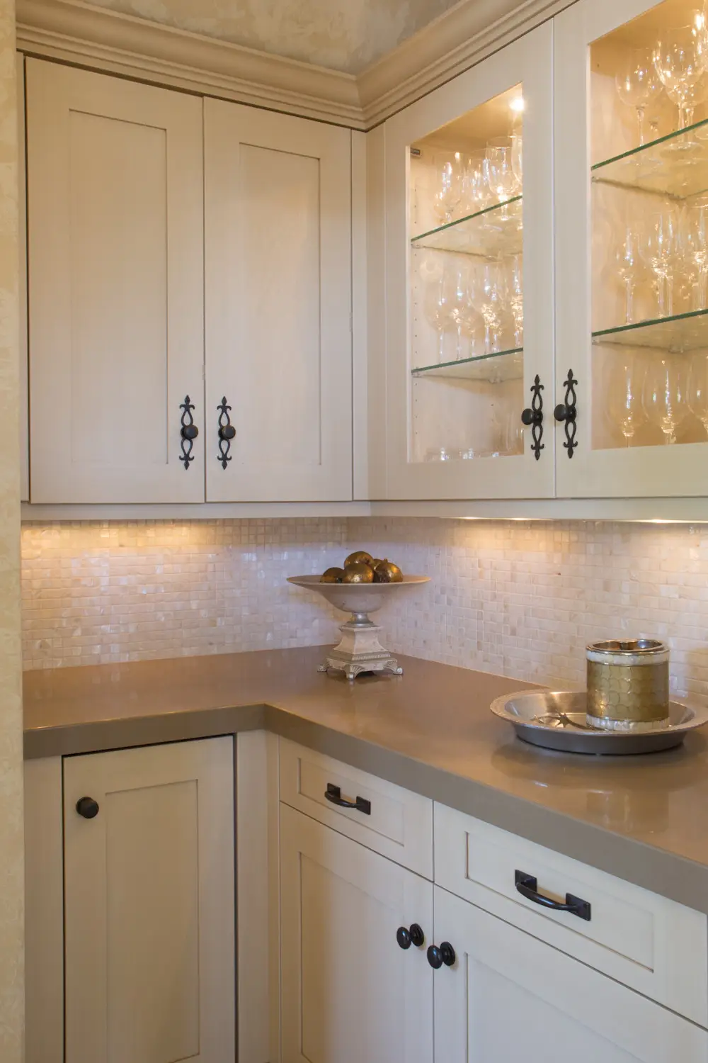 Elegant custom cabinetry in a Los Angeles home, featuring cream-colored cabinets with black iron handles, glass shelving displaying glassware, under-cabinet lighting, and a beige tiled backsplash, highlighting luxury storage solutions.
