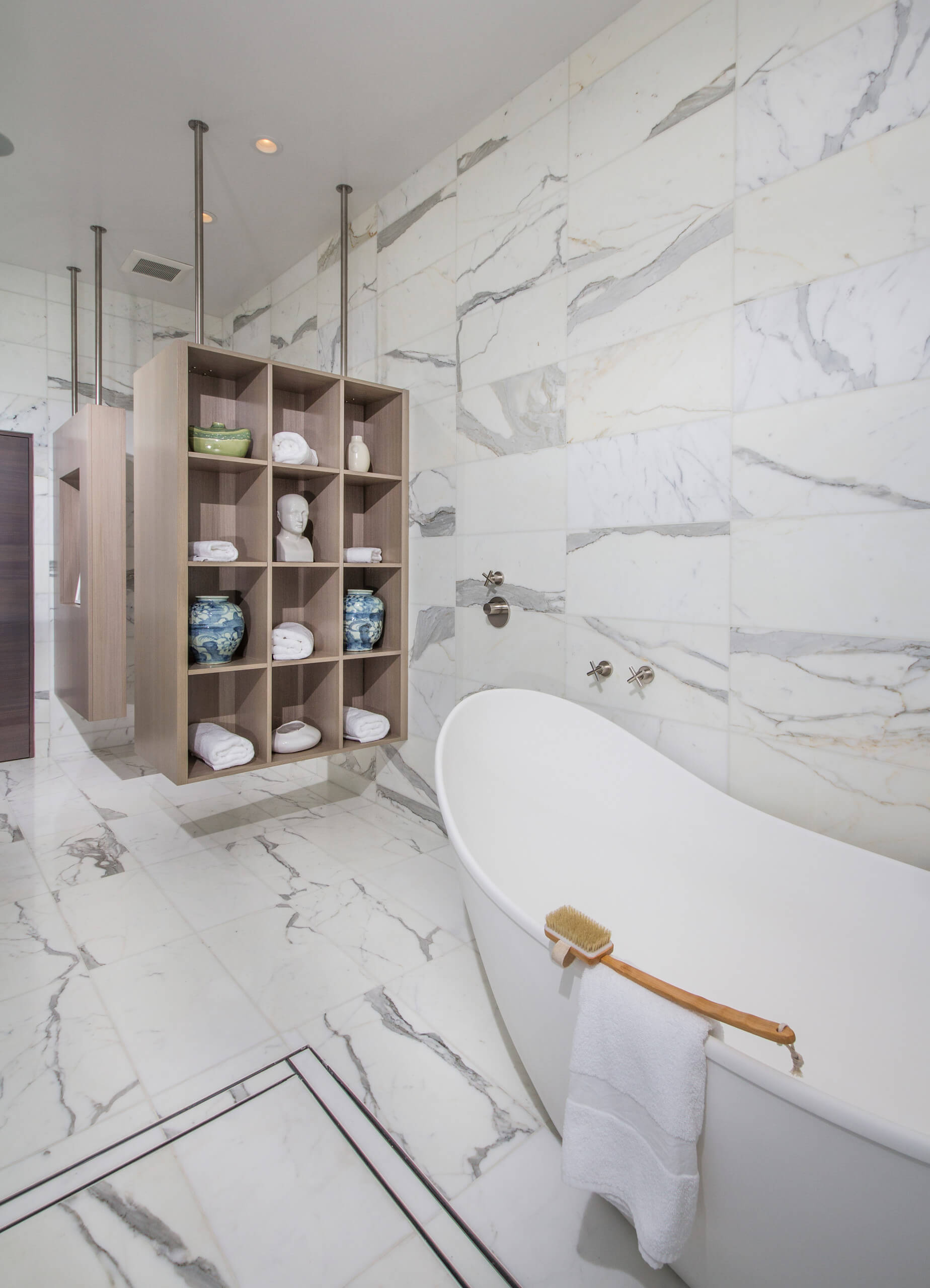 Luxurious bathroom with marble tiles, a freestanding white bathtub, and custom cabinets featuring storage for towels and decorative items. Premium custom cabinetry in Encinitas to enhance your space.