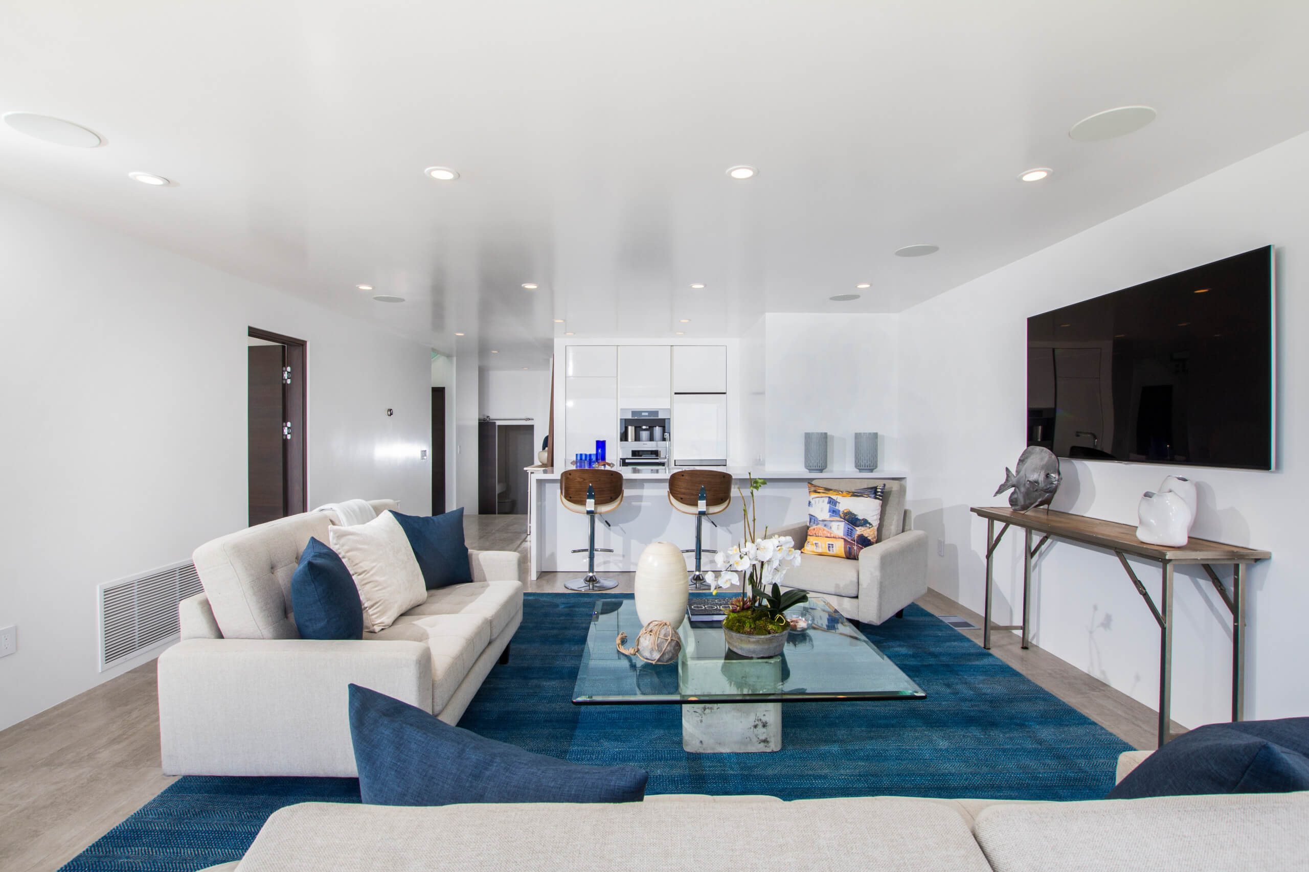 Modern living room with white custom cabinets in Encinitas, featuring a sleek design with a blue rug, glass coffee table, comfortable seating, and a large wall-mounted TV, offering a sophisticated space transformation.