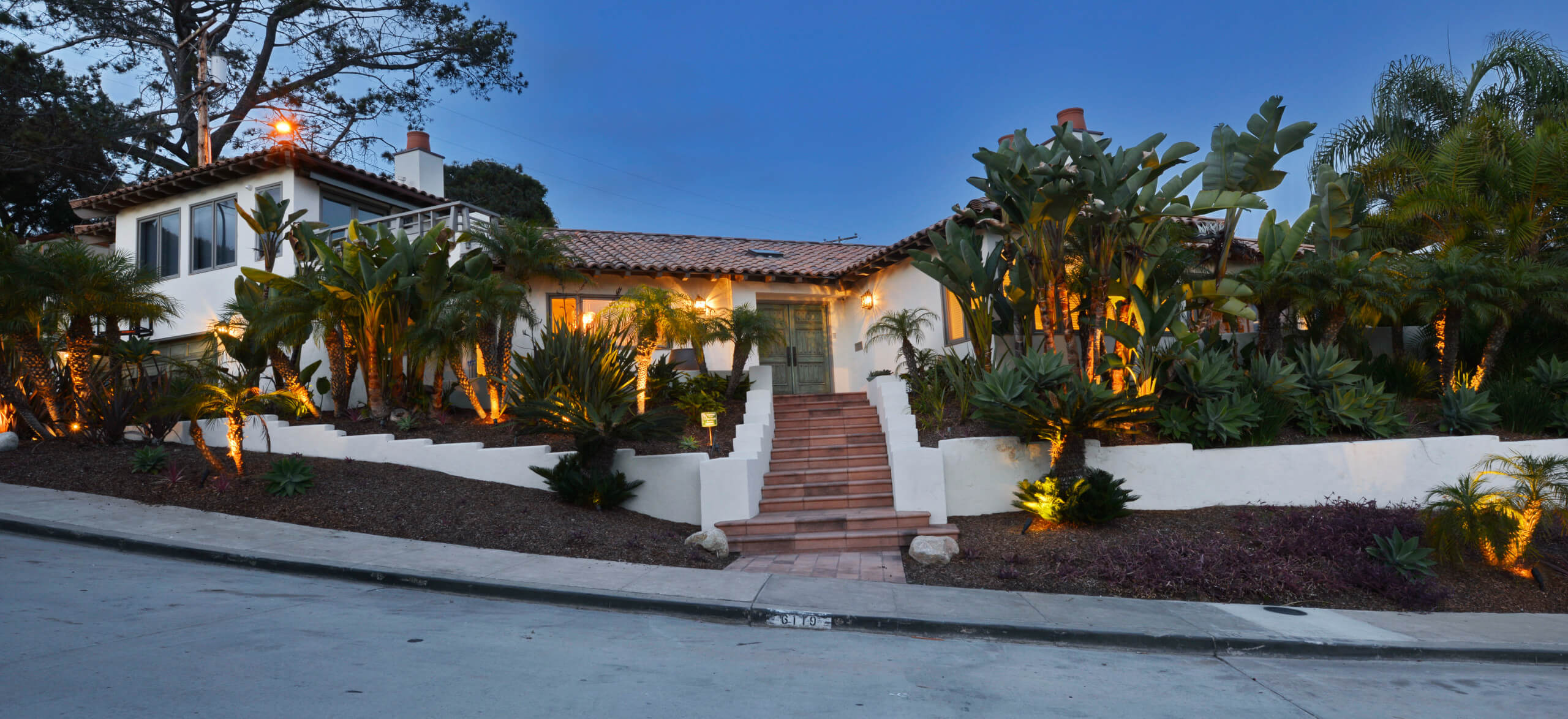 Elegant modern home exterior with lush landscaping and warm lighting, featuring a clay tile roof and wide steps leading to the front entrance.