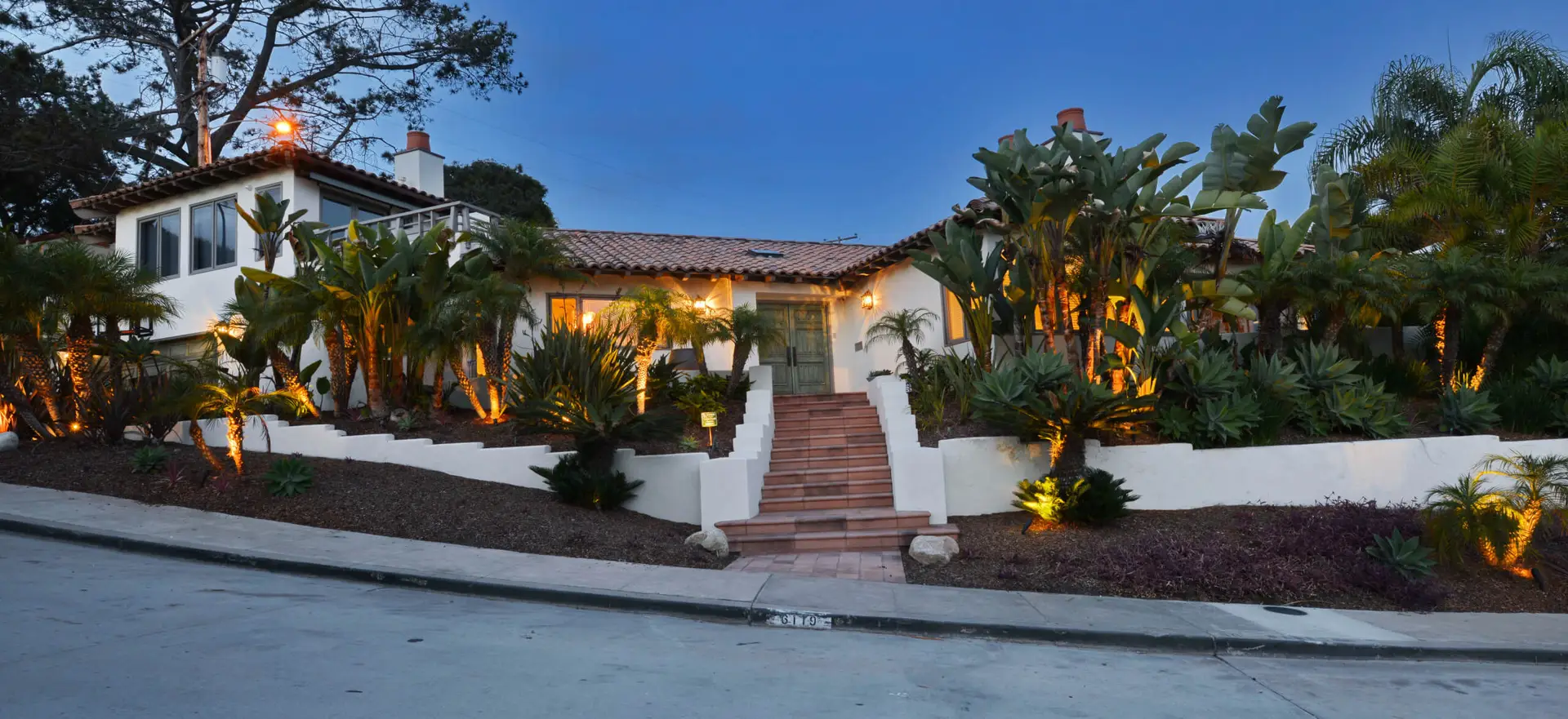 Elegant modern home exterior with lush landscaping and warm lighting, featuring a clay tile roof and wide steps leading to the front entrance.