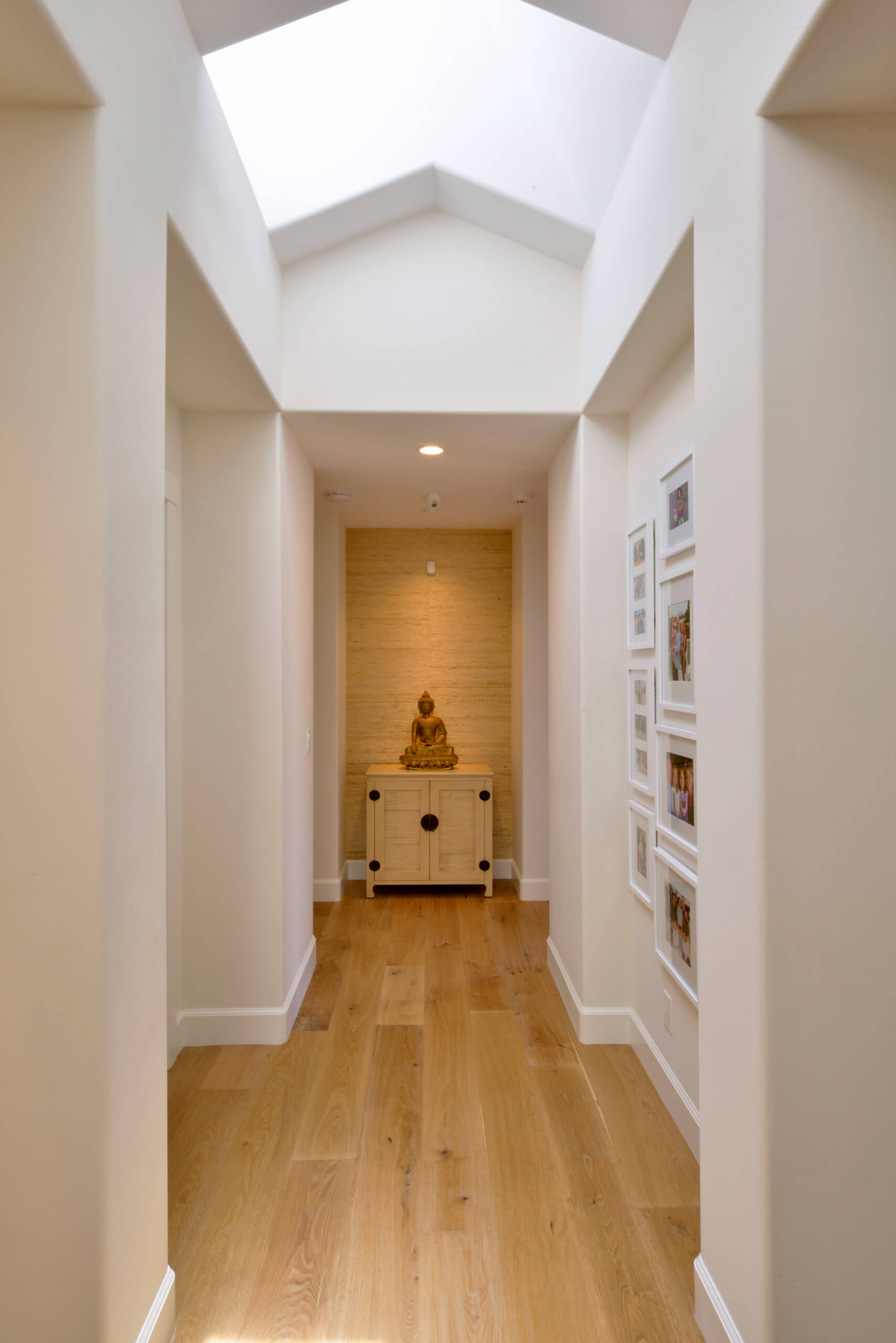 Modern home hallway with wooden floors and skylight, featuring a small cabinet with a Buddha statue, enhancing minimalist decor.