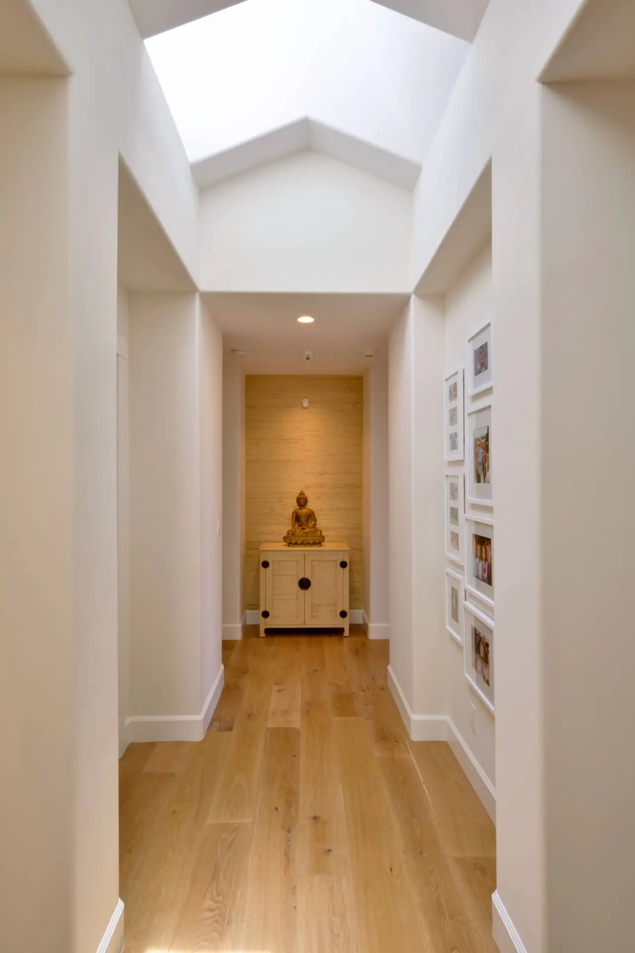 Modern home hallway with wooden floors and skylight, featuring a small cabinet with a Buddha statue, enhancing minimalist decor.