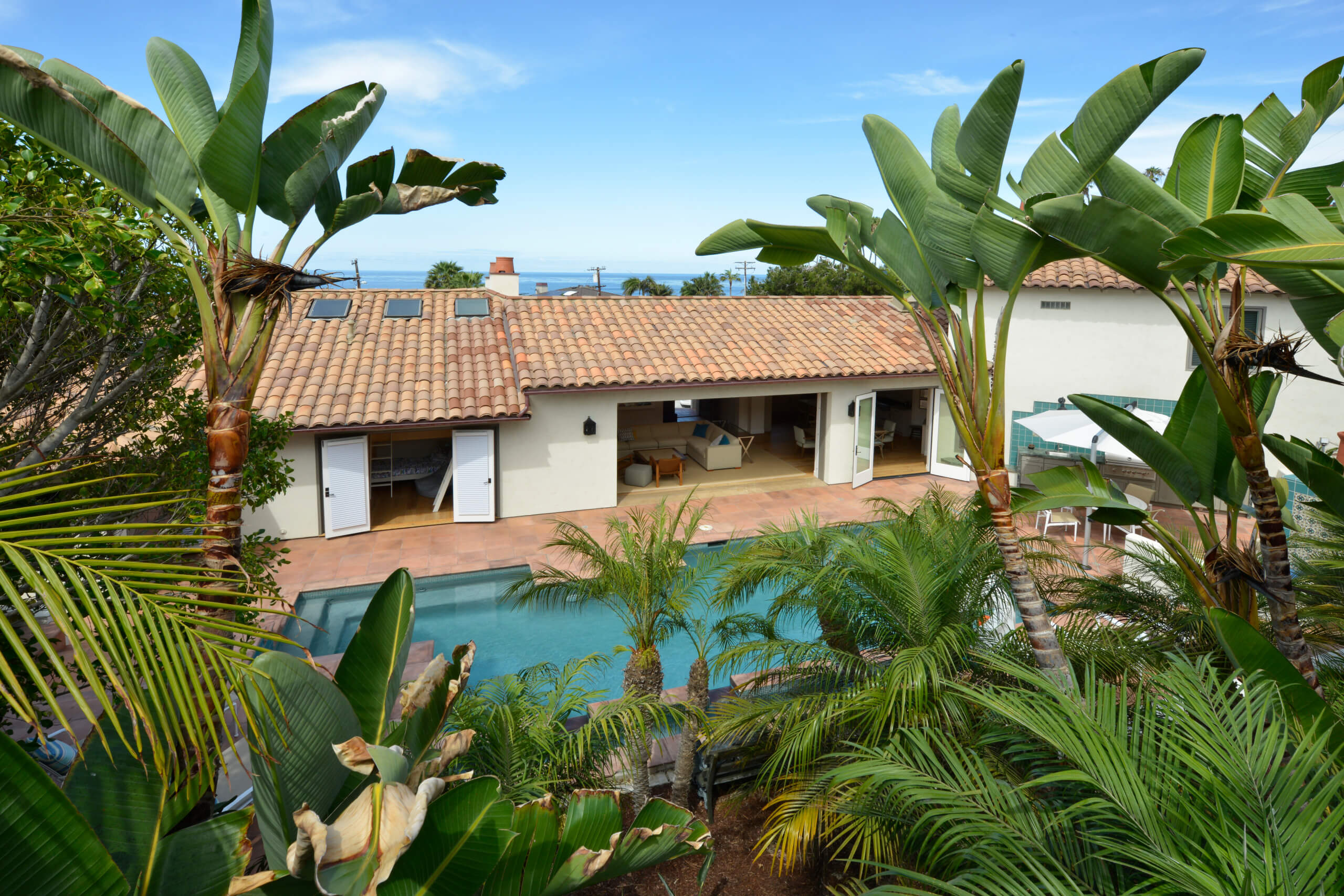 Modern home with tiled roof and open patio doors leading to a swimming pool, surrounded by lush palm trees, under a clear blue sky.