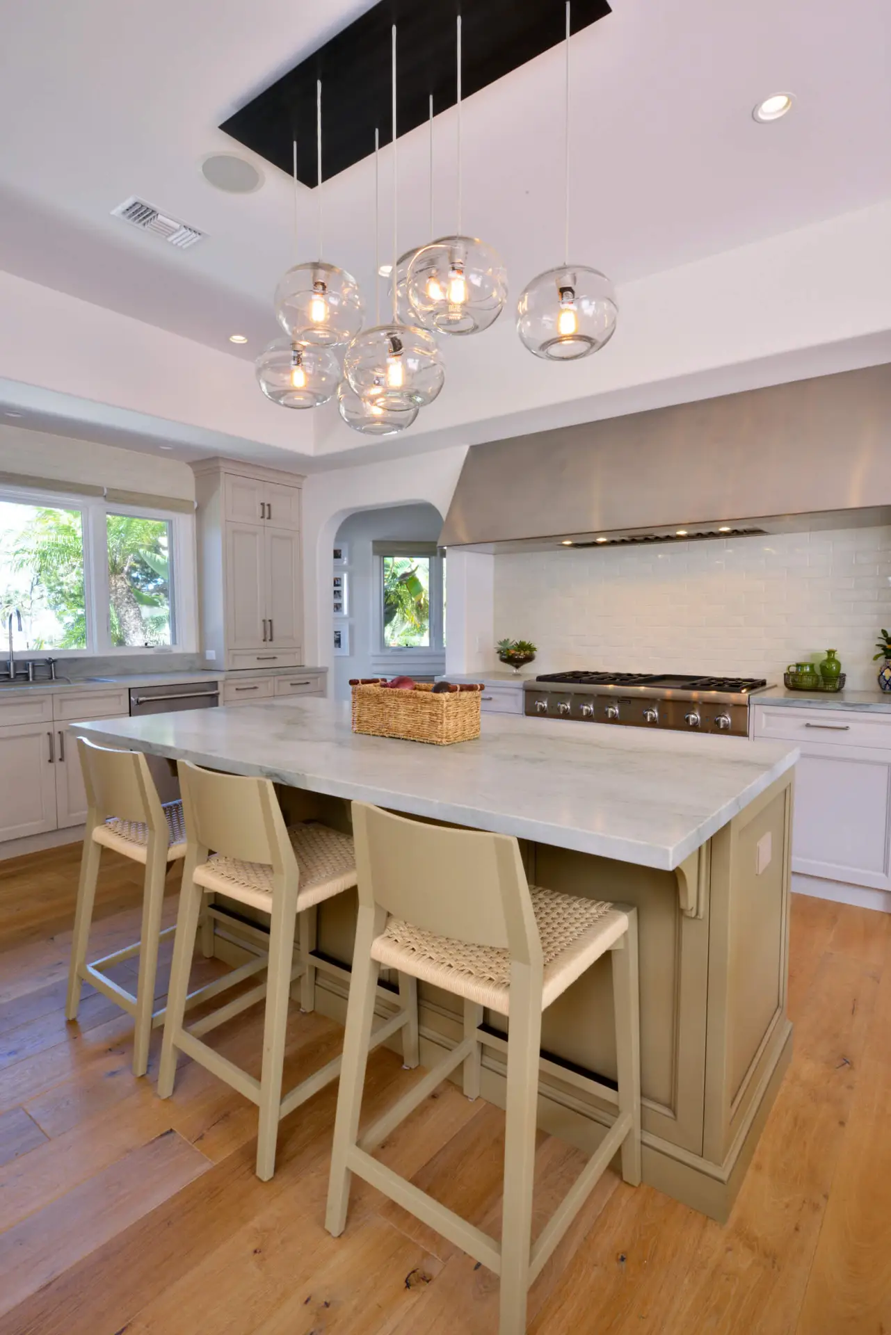 Modern kitchen with custom cabinets, a large central island featuring a marble countertop and wicker bar stools, contemporary pendant lighting, and a stainless-steel range hood. The room has natural light from a large window and wooden flooring.