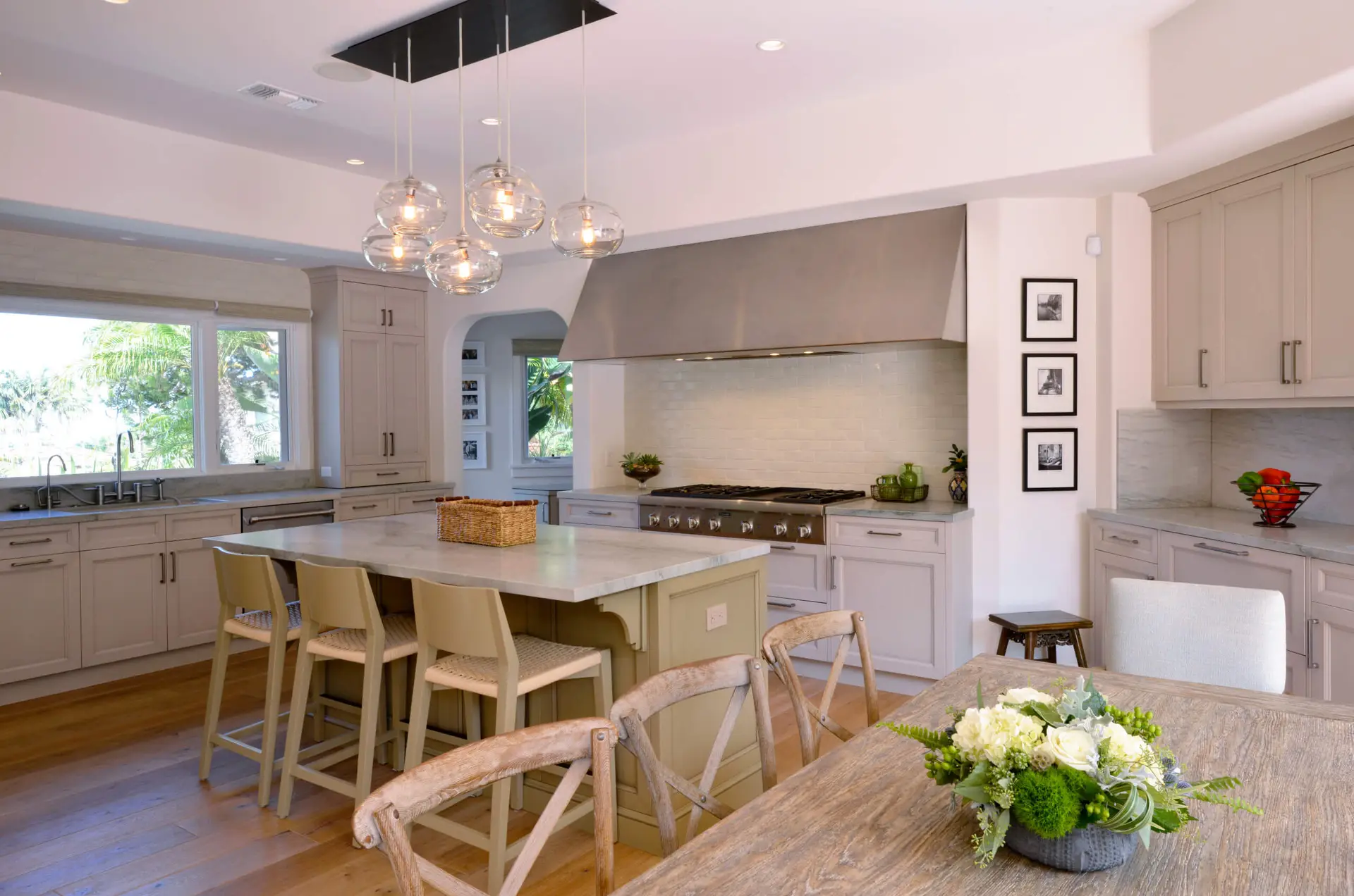 Modern kitchen with custom cabinets, marble island, and stylish lighting in a contemporary home. Features wooden floors, large windows, stainless steel appliances, and a floral centerpiece on a rustic dining table.