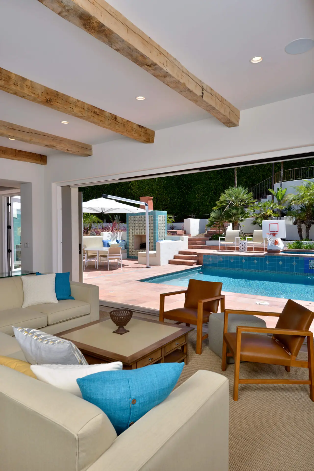 Modern living room with beige sofas and blue accent pillows opening to a stylish outdoor pool area. The space features wooden beams on the ceiling, outdoor seating, and lush greenery, highlighting luxury and seamless indoor-outdoor living.