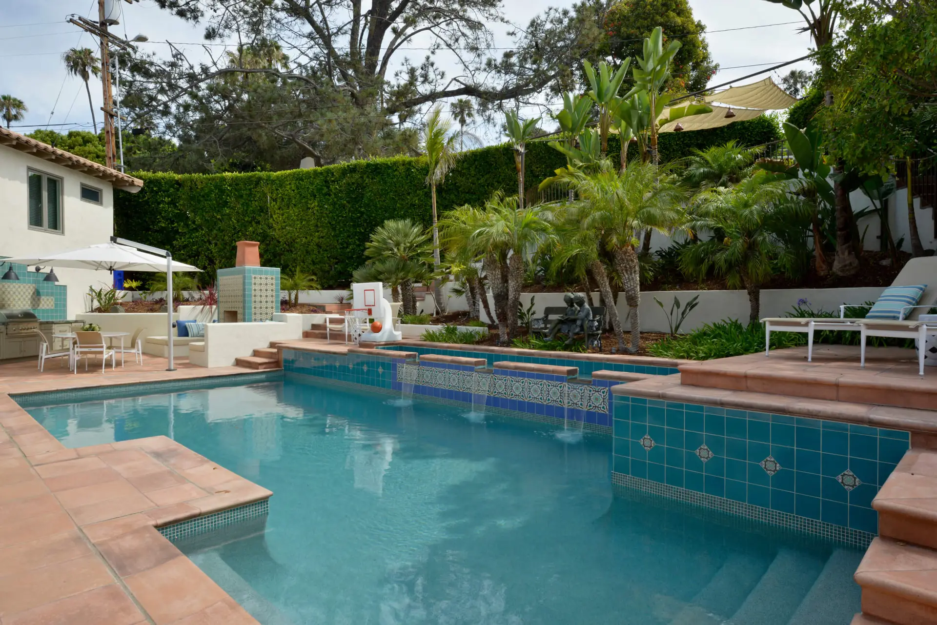 Spacious backyard featuring a luxurious swimming pool with turquoise tiles, surrounded by lush greenery and palm trees, with a stylish outdoor seating area and barbecue setup under large umbrellas.
