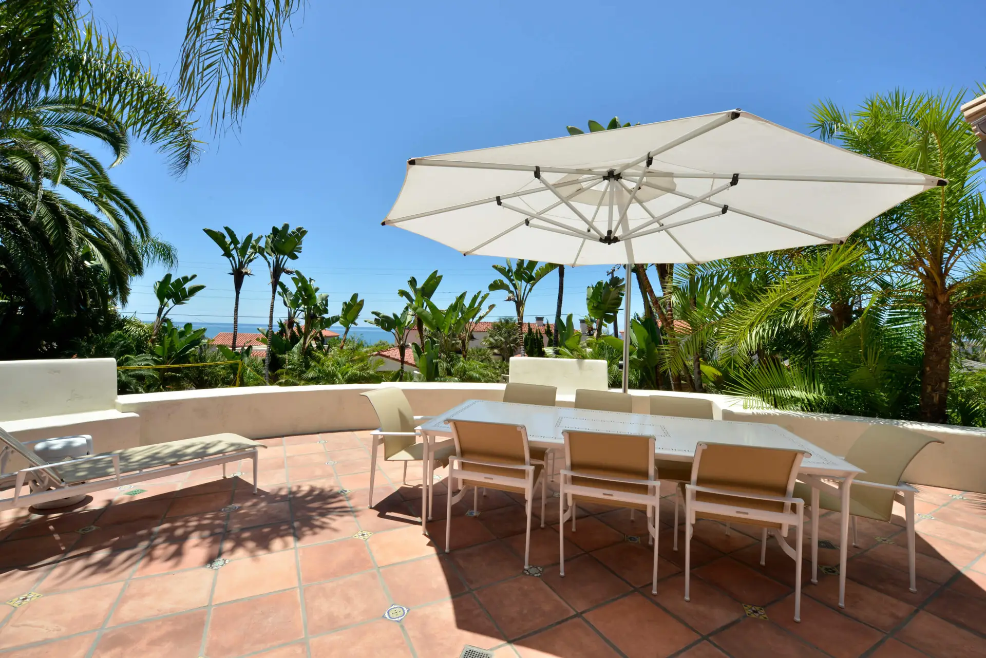 Sunny outdoor patio with a large white umbrella, modern dining table, chairs, and lush tropical plants. Perfect for relaxing in a modern home environment.