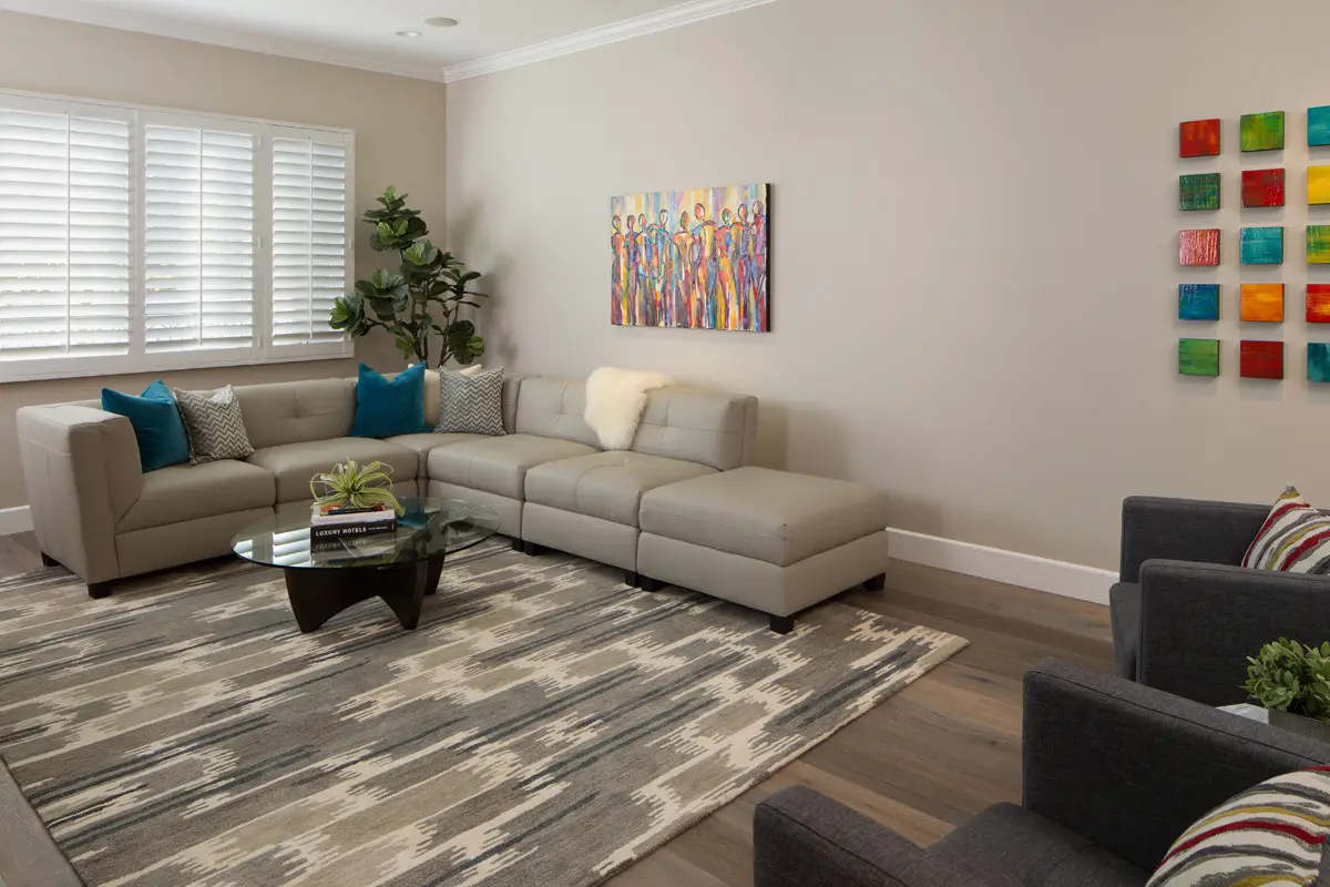 Interior of a modern living room with custom cabinetry, featuring a beige sectional sofa with colorful pillows, a glass coffee table on a patterned rug, contemporary artwork on the walls, and a large window with white shutters.