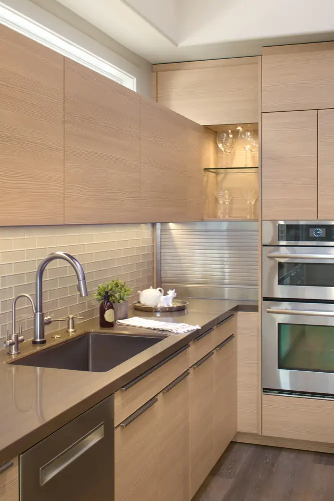 Modern kitchen featuring custom wood cabinetry with a sleek design. The brown cabinets are complemented by a dark countertop, stainless steel fixtures, and an integrated oven. Overhead cabinets include a section with glass doors showcasing glassware. The backsplash is made