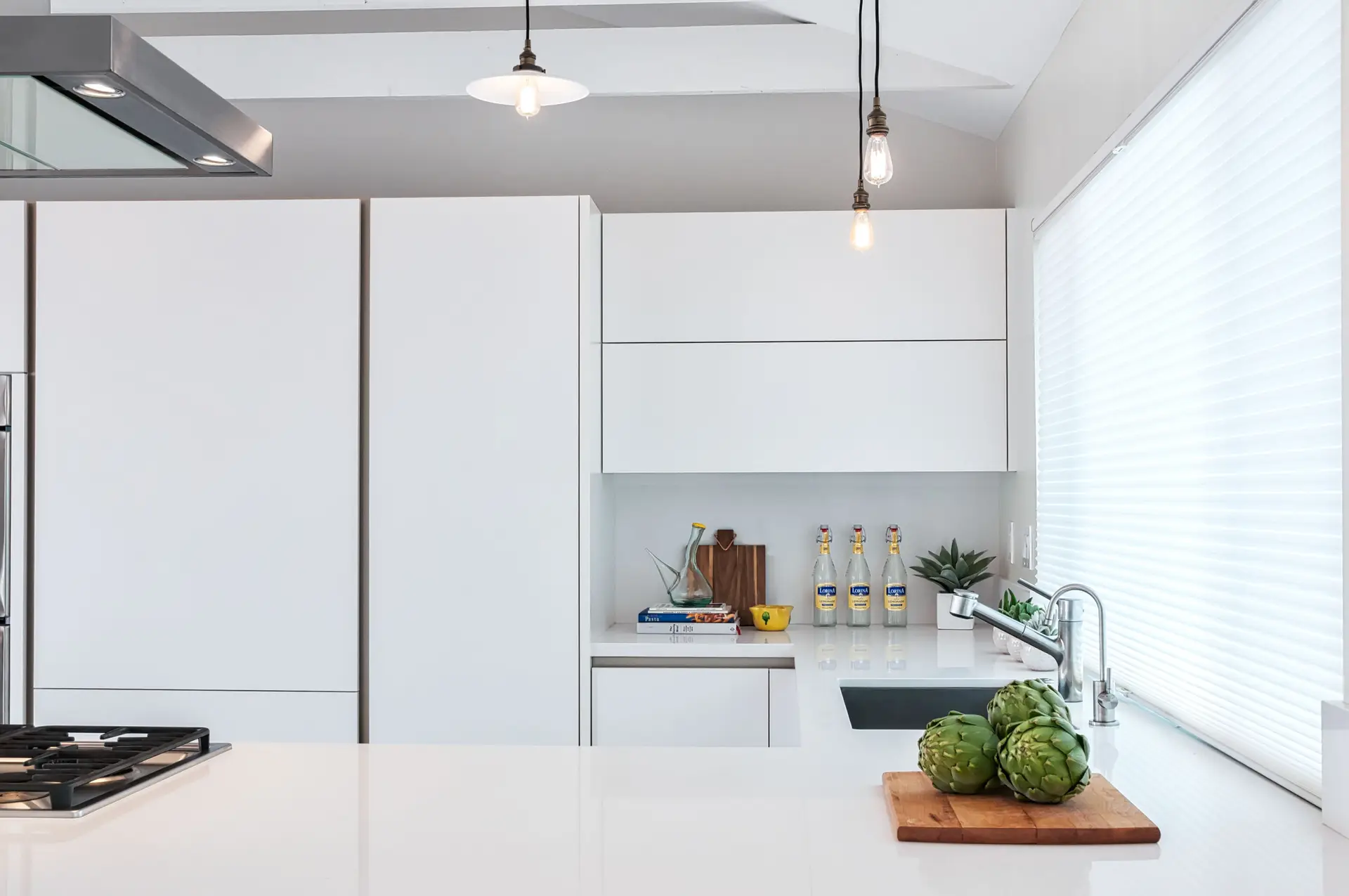 Modern luxury kitchen design featuring sleek white cabinets and a minimalist decor. Bright natural light fills the space, highlighting elegant features like a chrome faucet, artichokes on a cutting board, and stylish pendant lighting. Perfect for creating an elegant and sophisticated atmosphere.