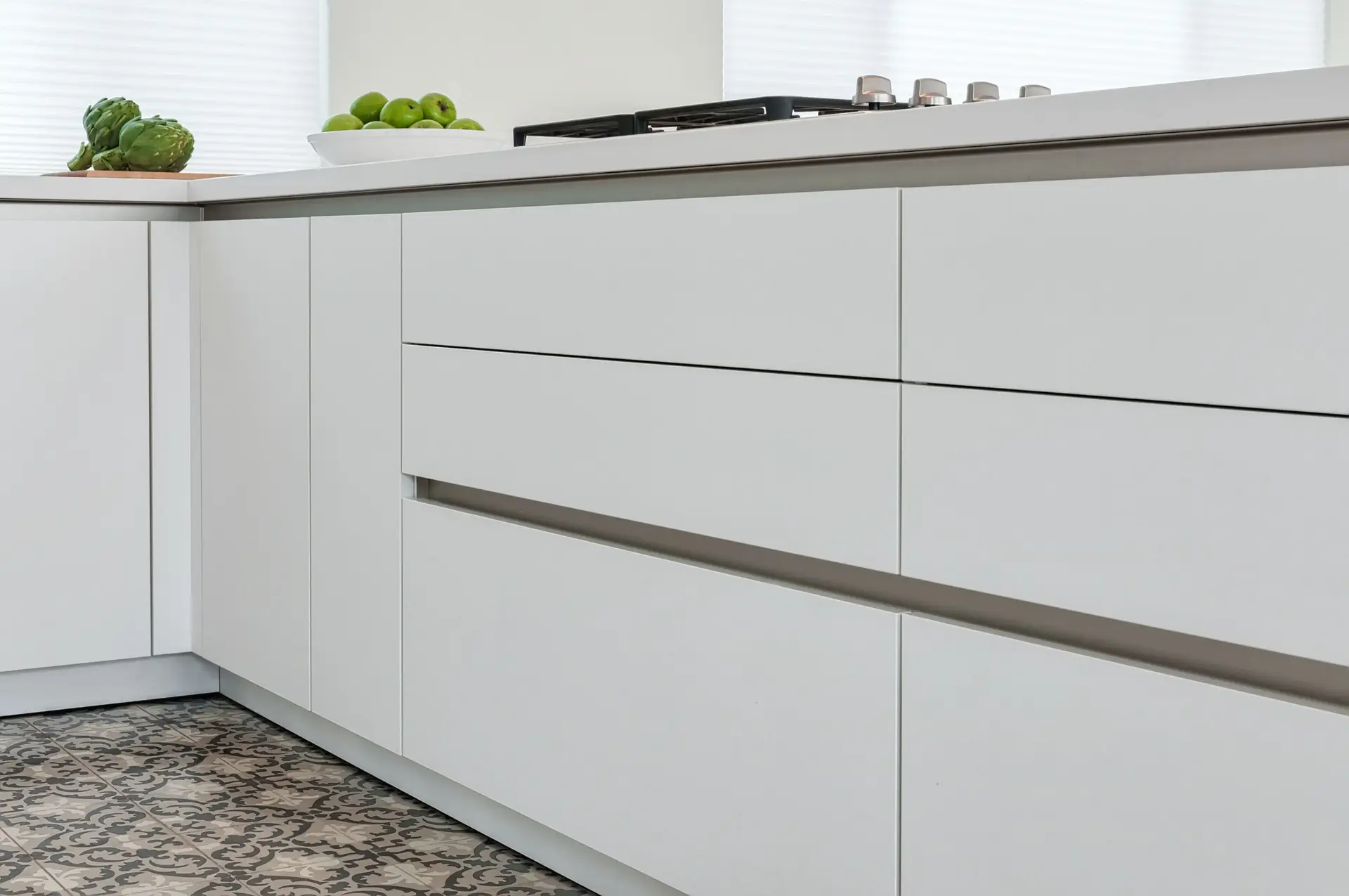 Modern luxury kitchen design featuring sleek white cabinets with minimalist handles, elegant patterned flooring, and a countertop adorned with fresh green apples and artichokes for a sophisticated look.
