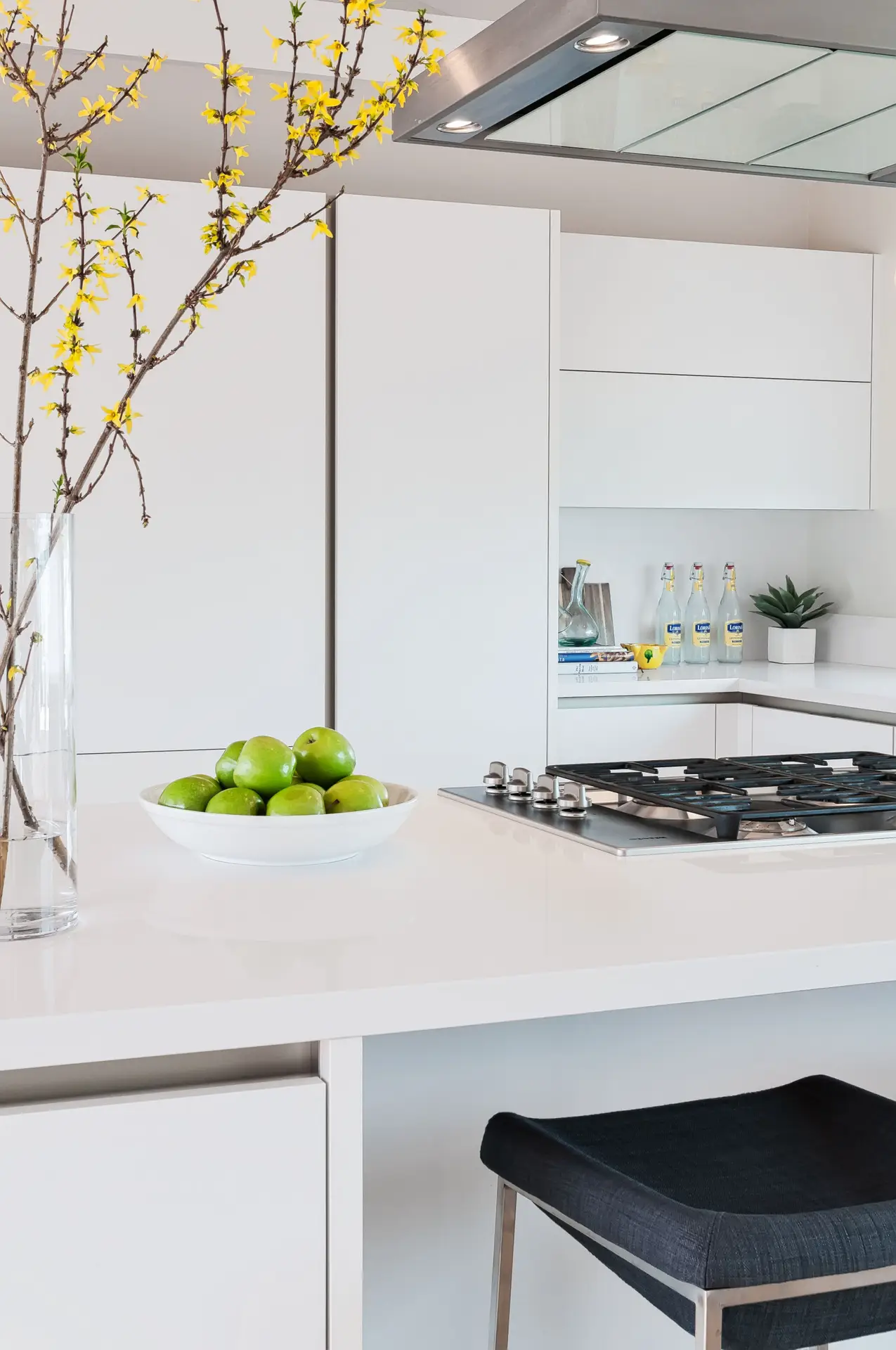 Modern luxury kitchen with sleek white cabinets, minimalist design, and elegant decor. A bowl of green apples sits on the countertop, complemented by a vase with yellow flowers. A gas cooktop is integrated into the countertop, adding functionality to the stylish space.