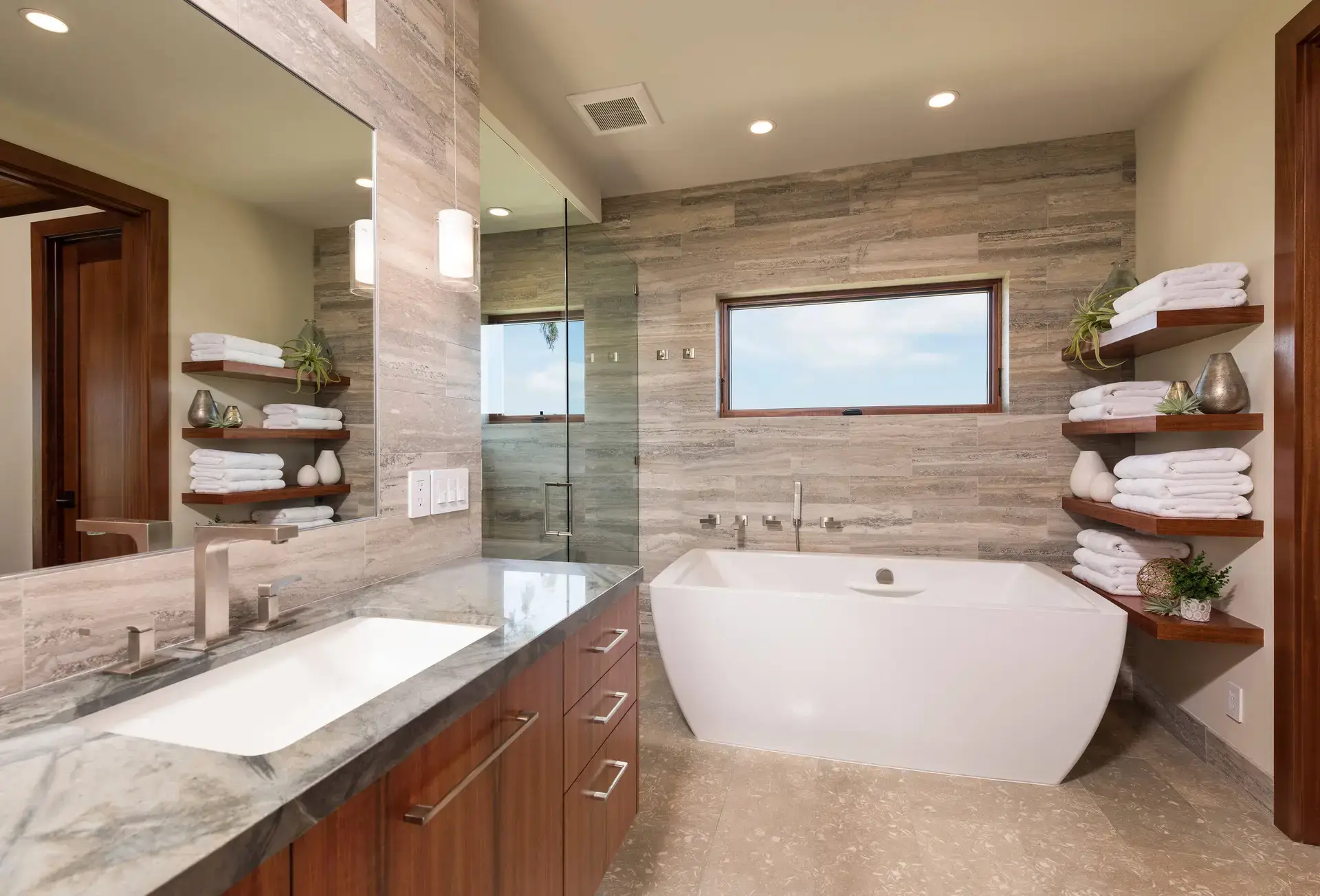 Luxurious bathroom featuring custom wooden cabinetry, elegant stone countertops, and modern fixtures in San Diego & LA. Contemporary design with a freestanding bathtub and wall shelves displaying neatly stacked towels.