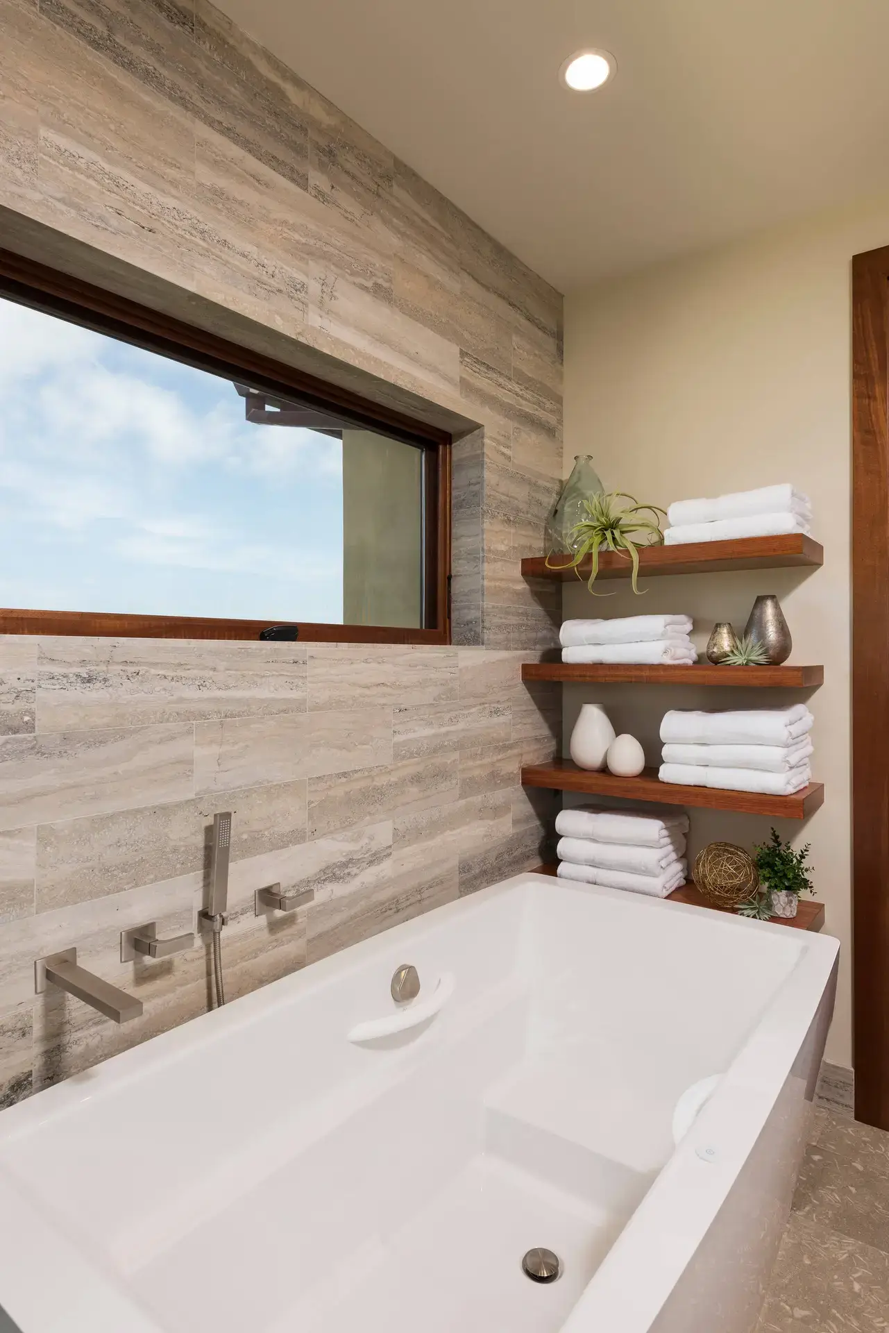 Luxurious bathroom with a modern white bathtub, elegant wooden shelves holding neatly folded towels and decorative items, and a large window providing natural light. High-end bathroom design in San Diego and LA.